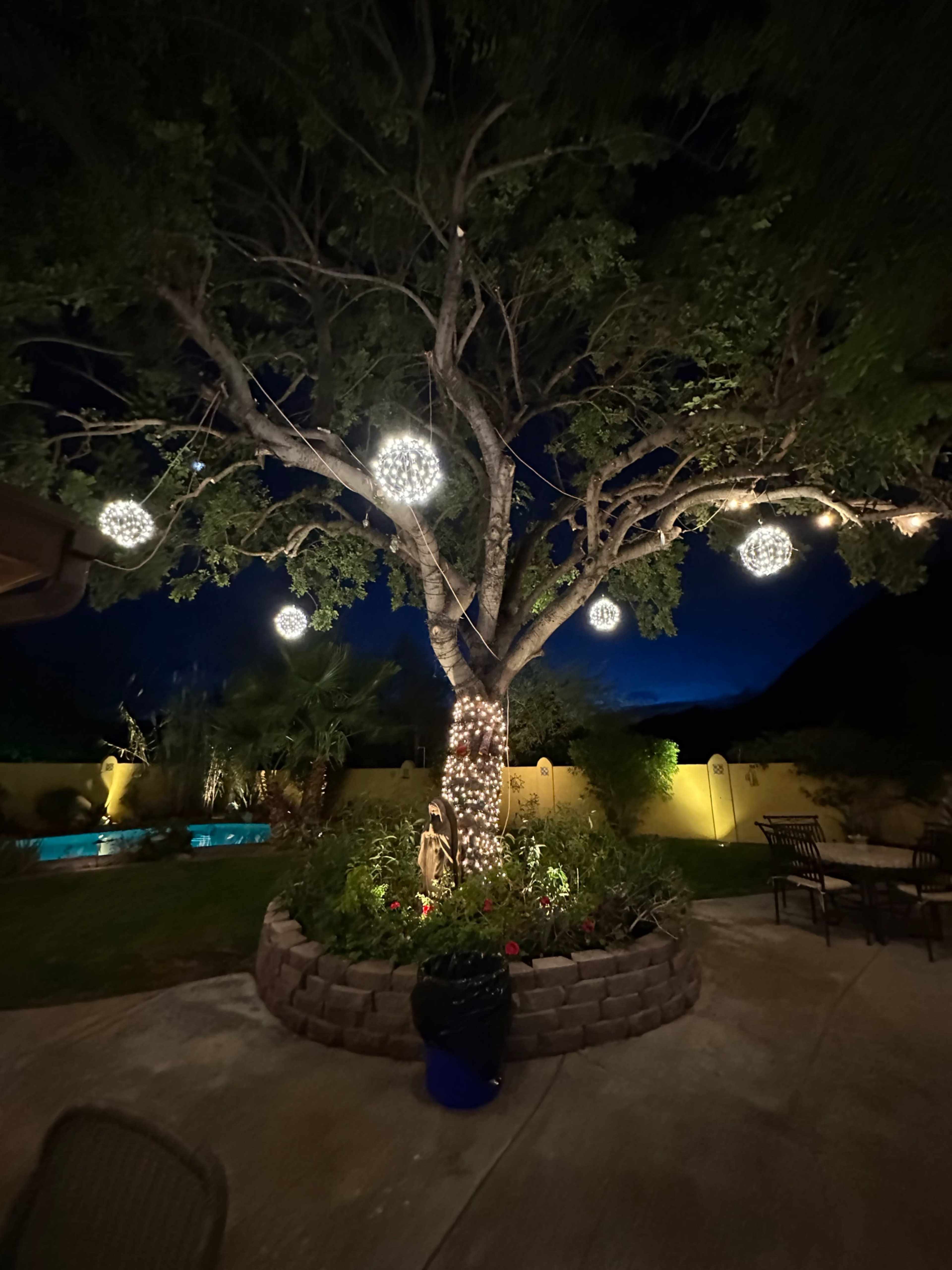 A tree adorned with round lights stands in a backyard at night, surrounded by a stone planter and a small swimming pool in the background.