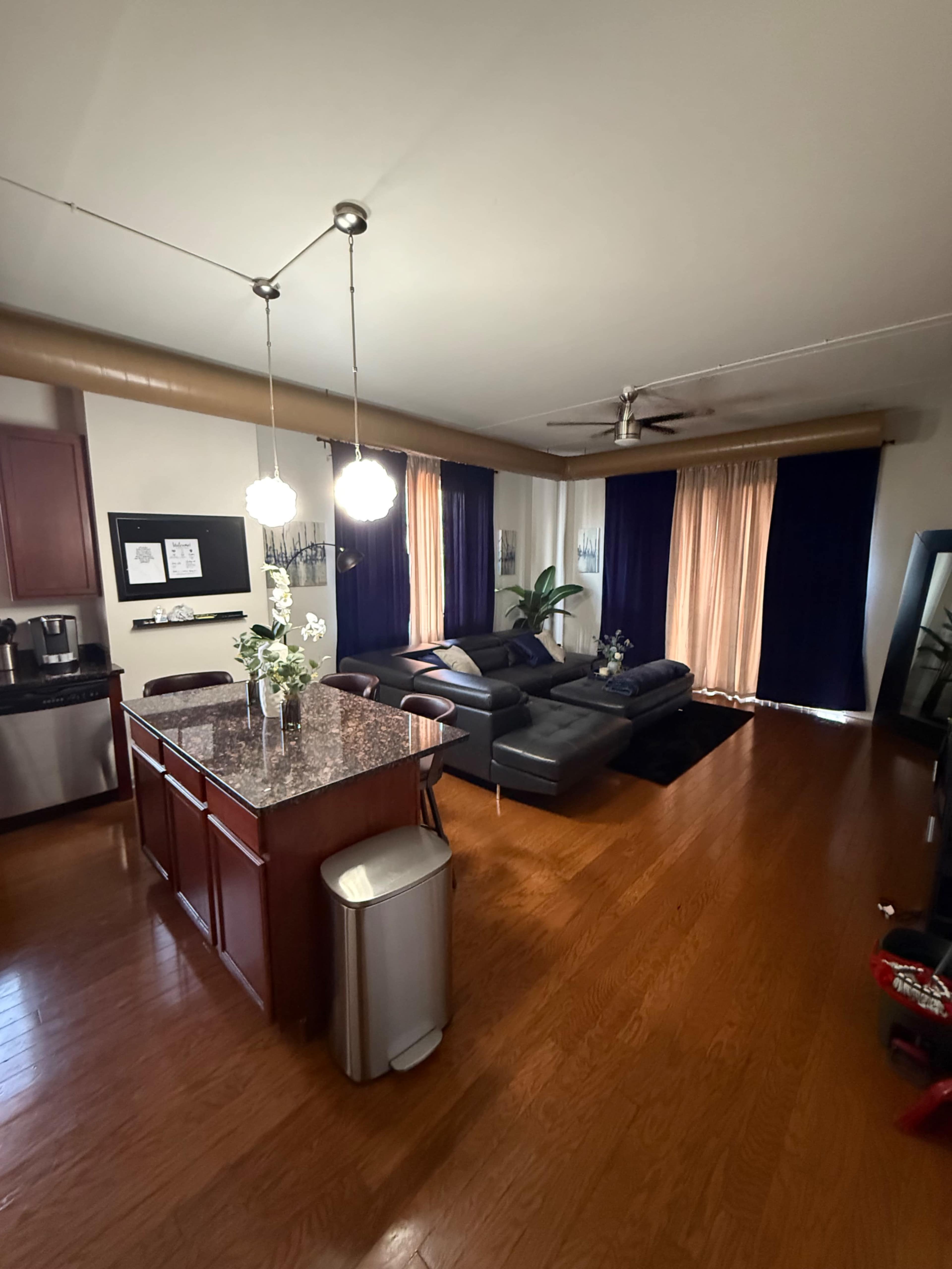 The image shows a spacious living area with a kitchen island, modern sofas, and large windows covered by sheer and dark curtains.