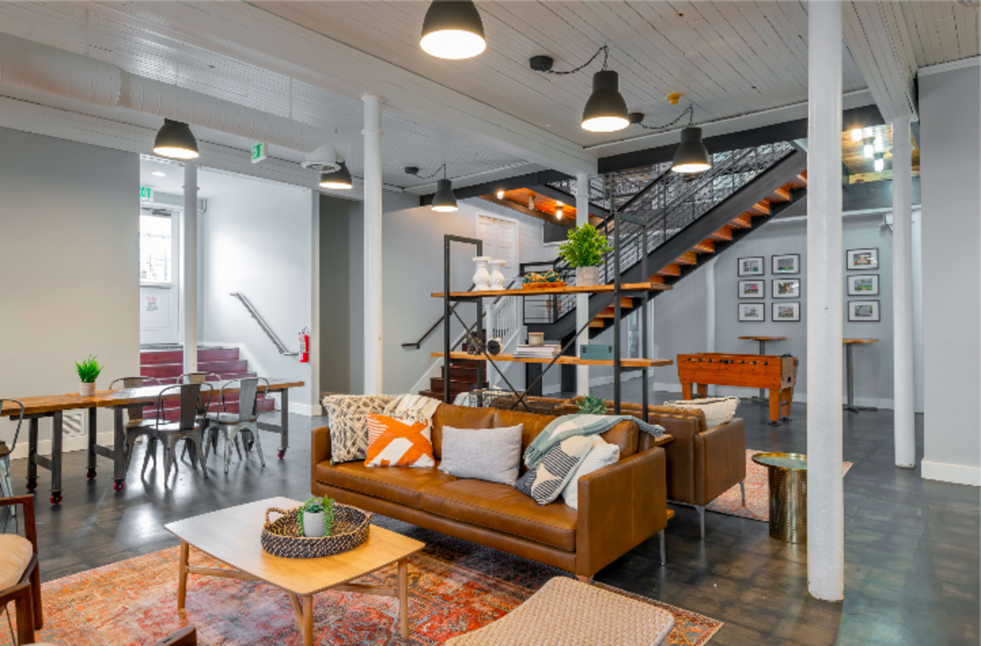 A modern, open-plan communal space featuring a brown leather sofa, wooden coffee tables, and a staircase leading to an upper level, with a mix of seating areas and decor elements.