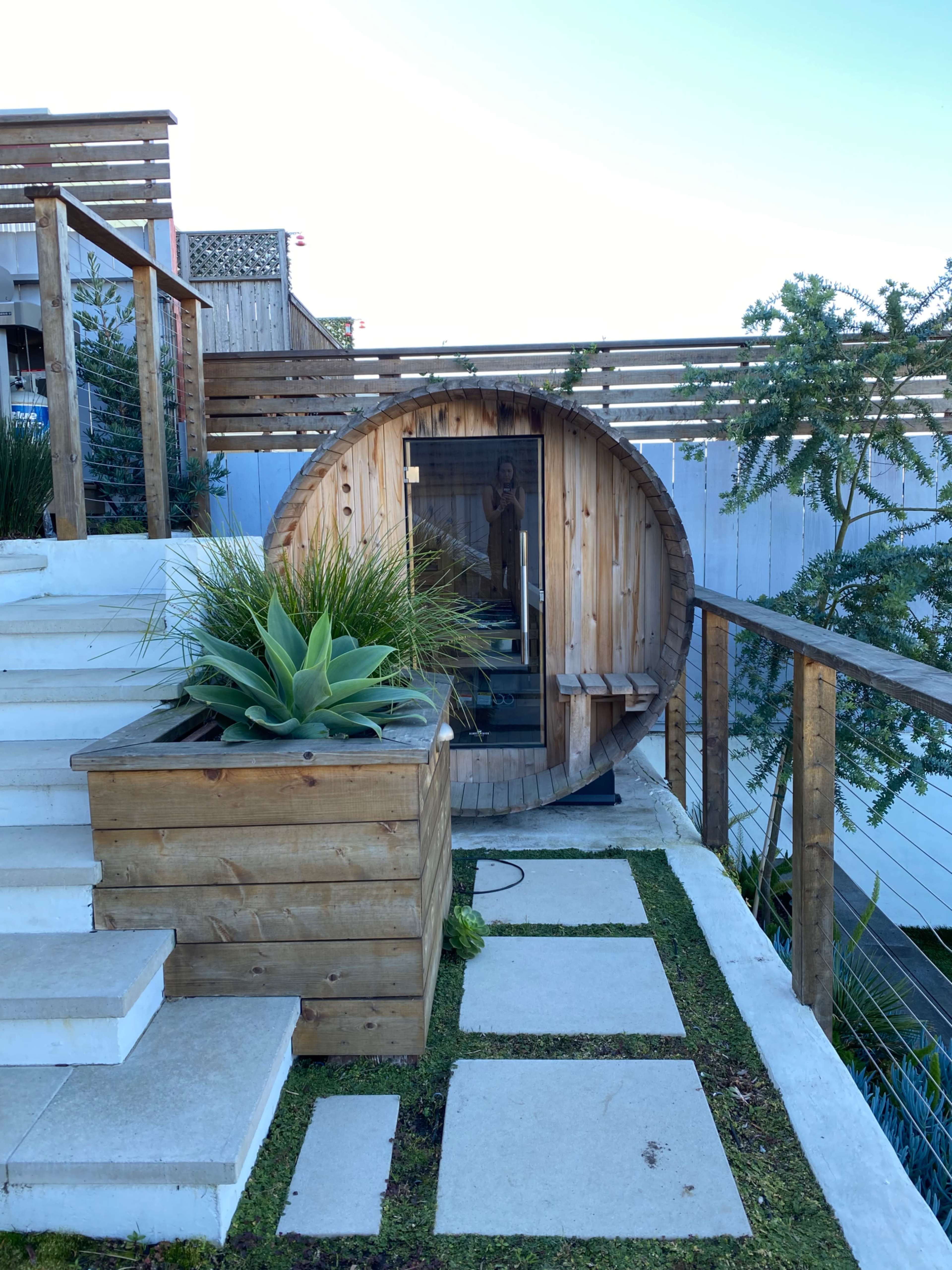 A wooden barrel sauna is positioned next to stone steps and a planter featuring tall grass and succulents.
