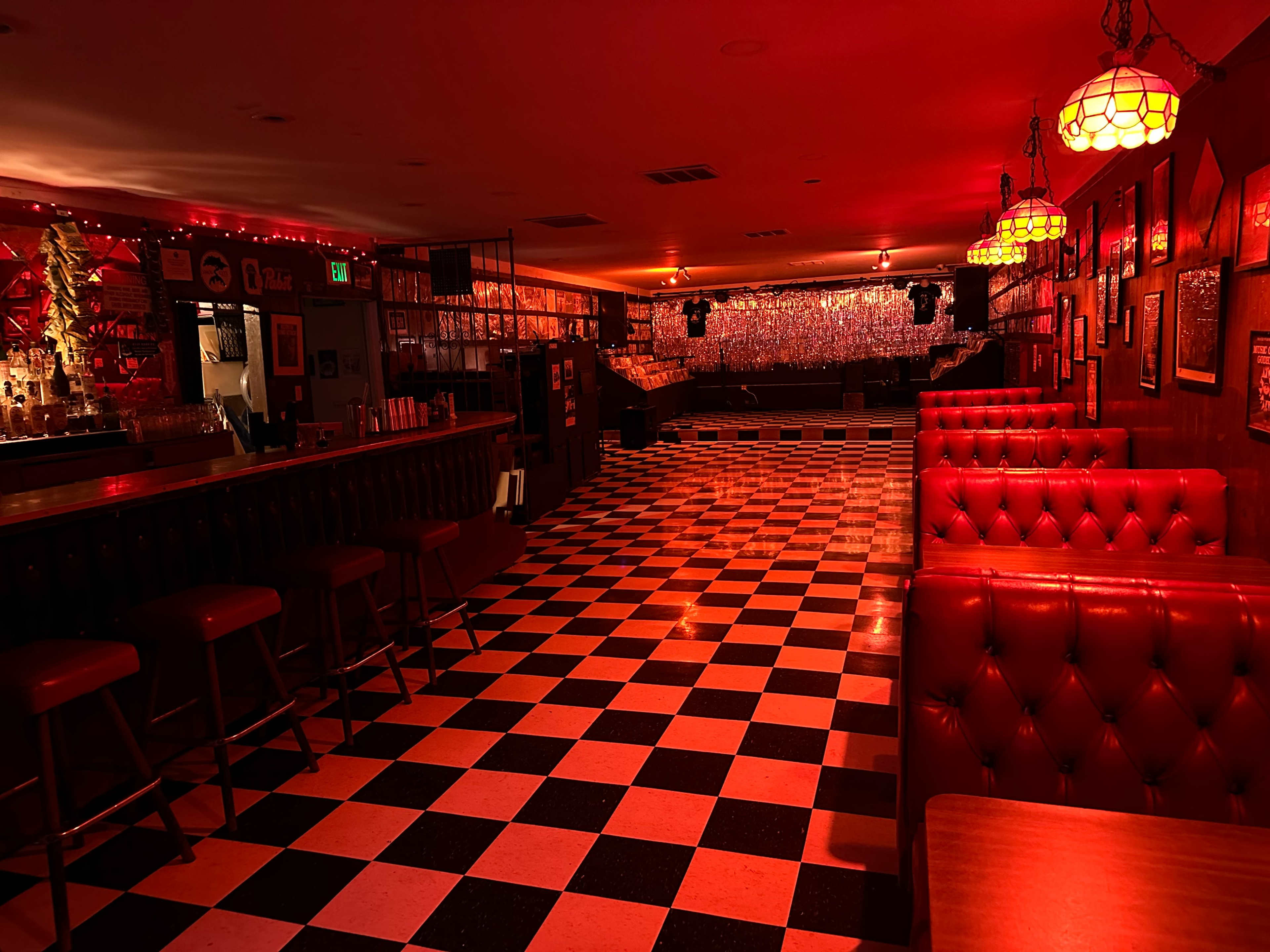 The image shows a dimly lit bar with a red color scheme, featuring a checkerboard floor and a long counter lined with stools.