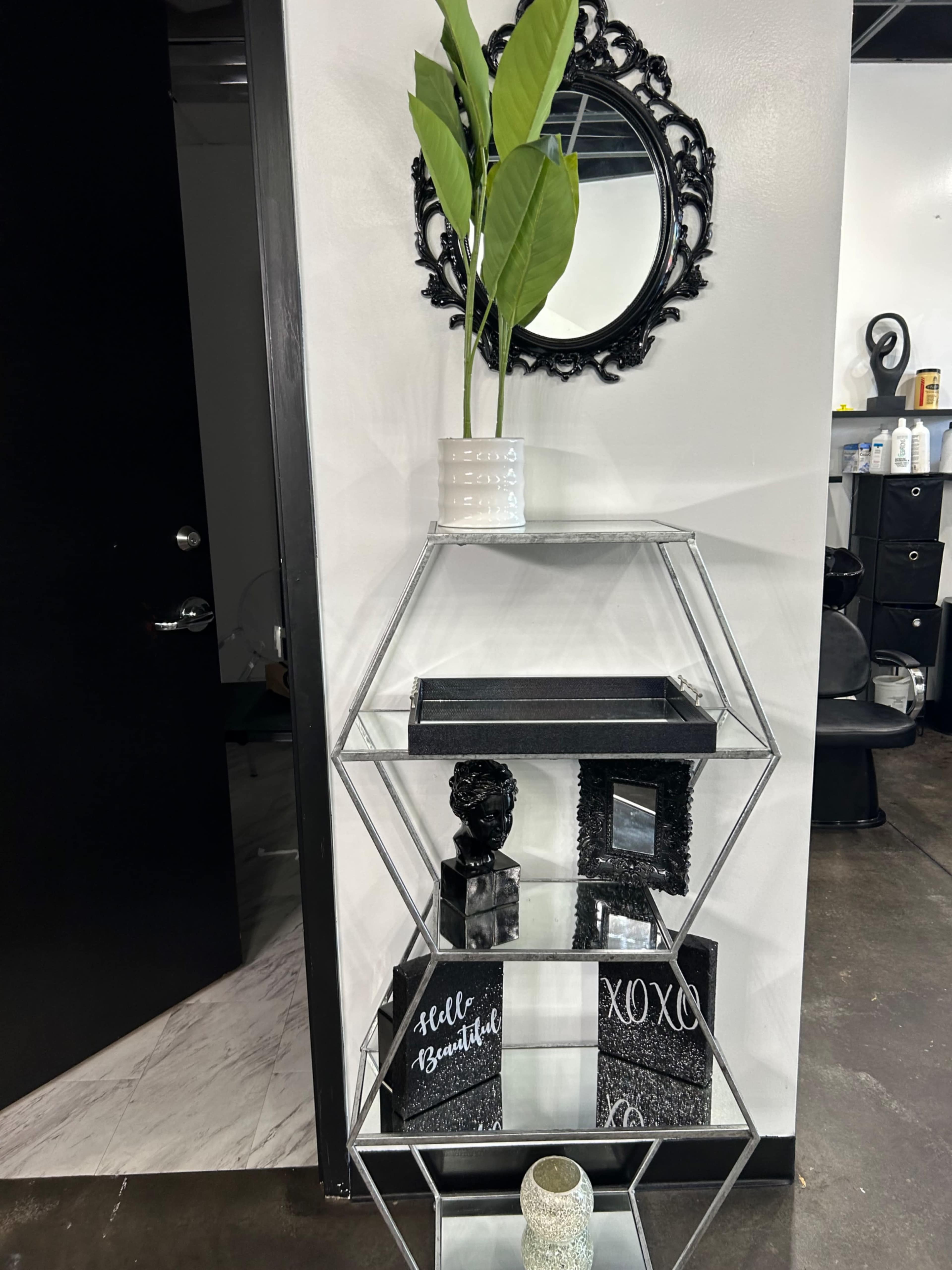 A metallic shelf with angular tiers displays a potted plant, a decorative bust, and various artistic signs against a contrasting black wall.