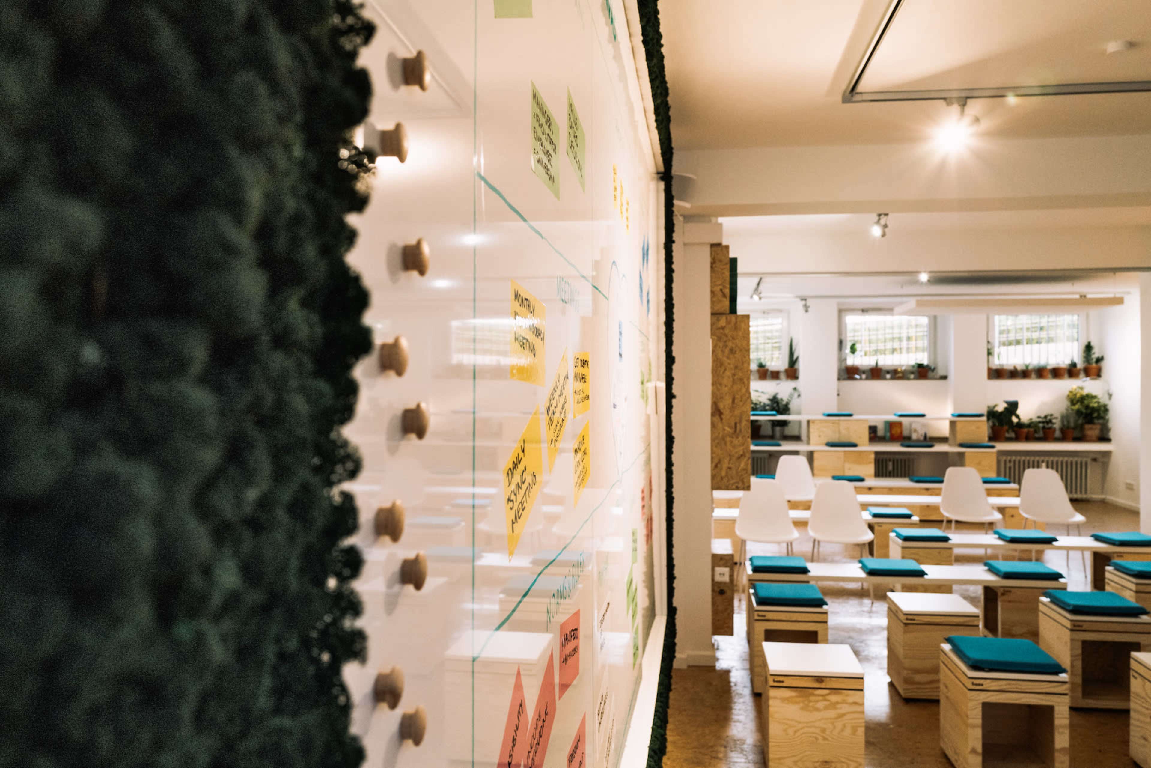 The image shows a spacious room equipped with wooden seating and a collaborative wall filled with colorful notes, surrounded by greenery.