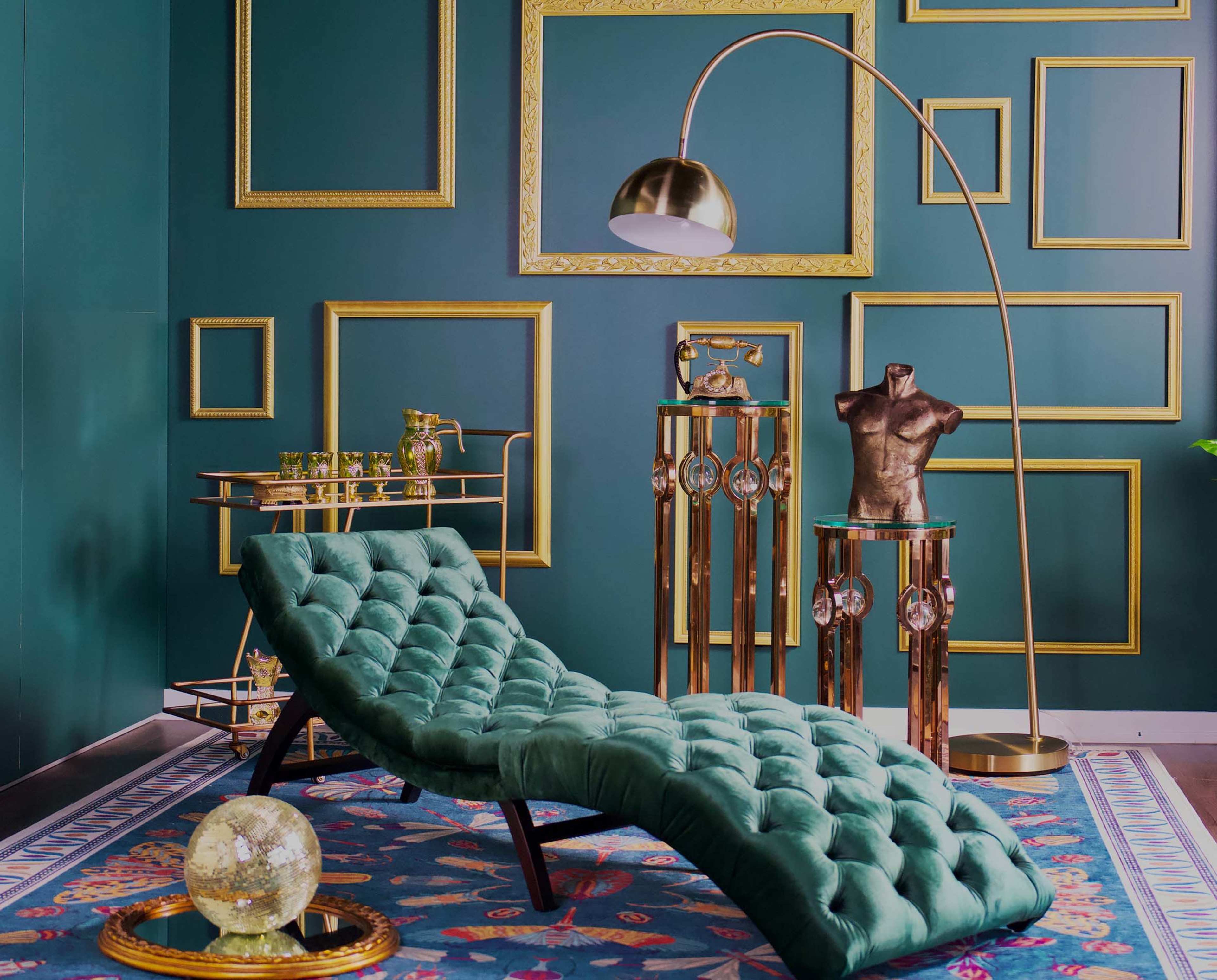 The image features a green tufted chaise lounge in a room with dark green walls adorned with various empty golden picture frames, a gold floor lamp, and decorative furniture.