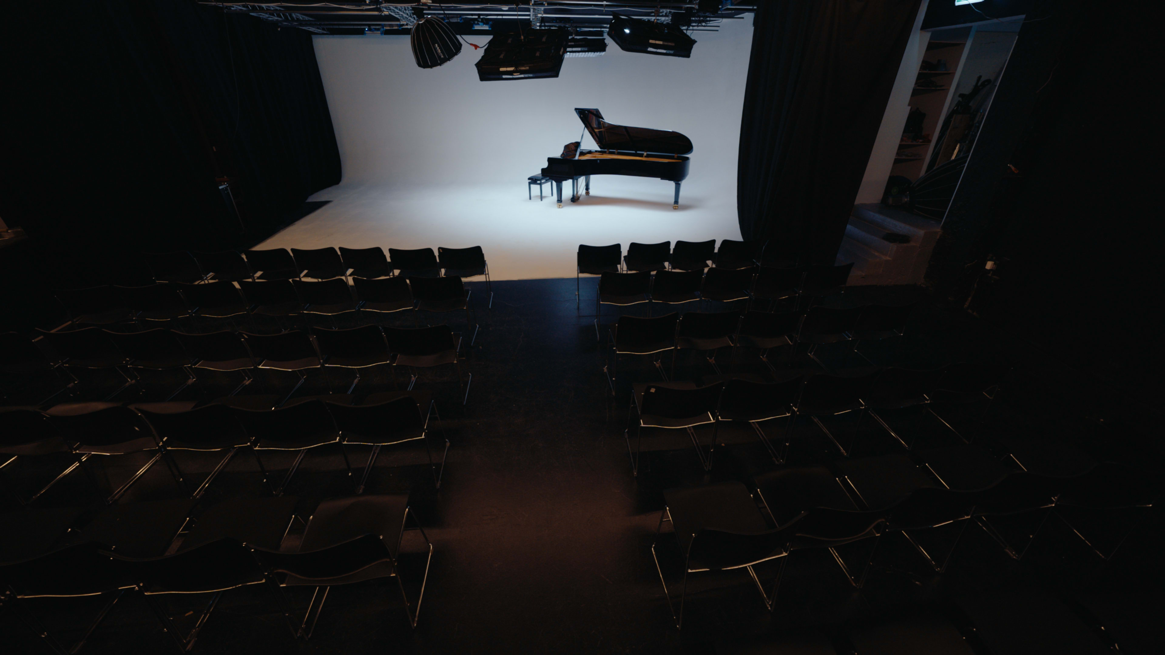 The image shows an empty performance space with rows of black chairs facing a grand piano on a white stage surrounded by dark curtains.