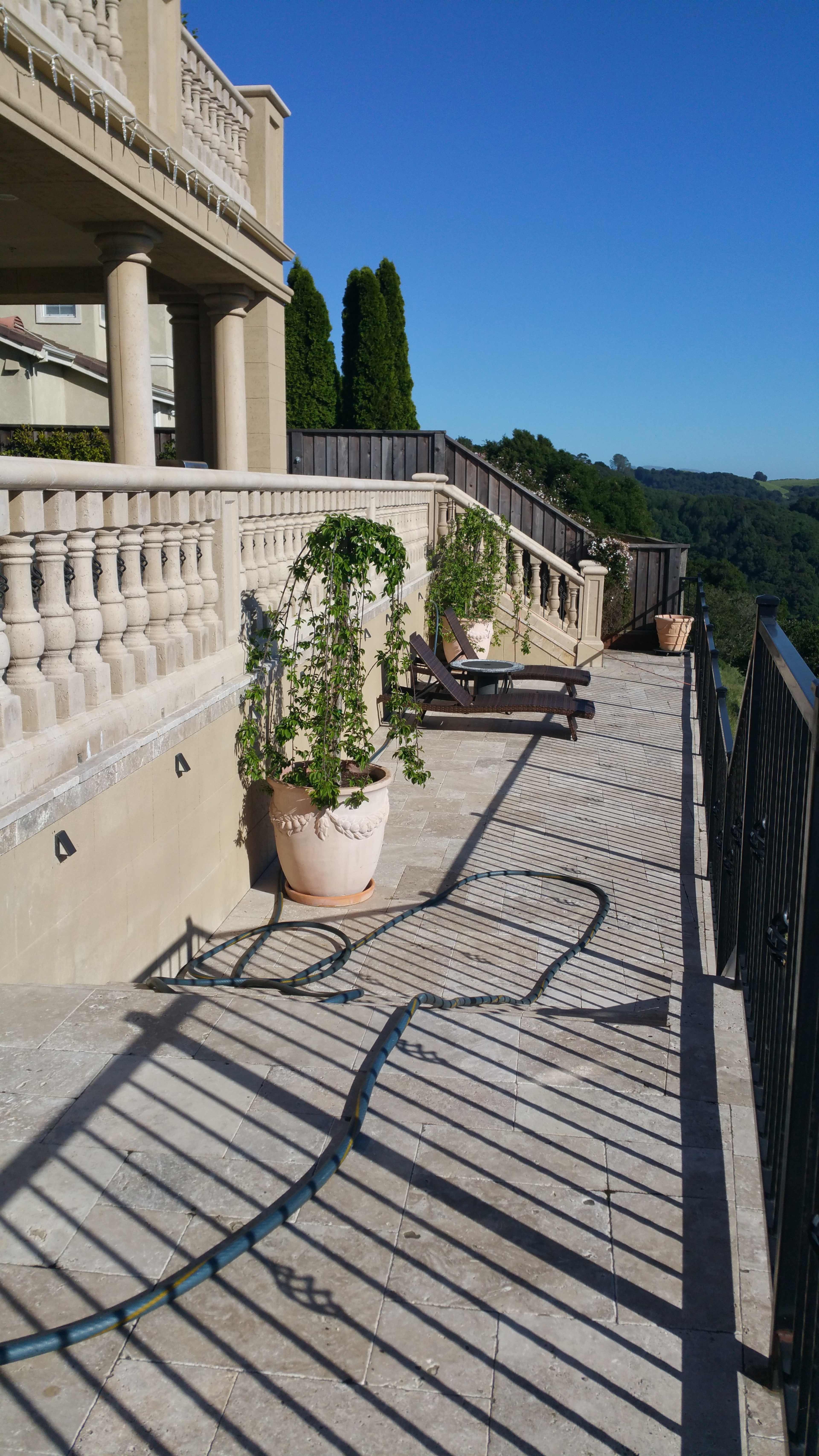 A stone terrace with railings, potted plants, a lounge chair, and a green garden hose partially coiled along the ground.