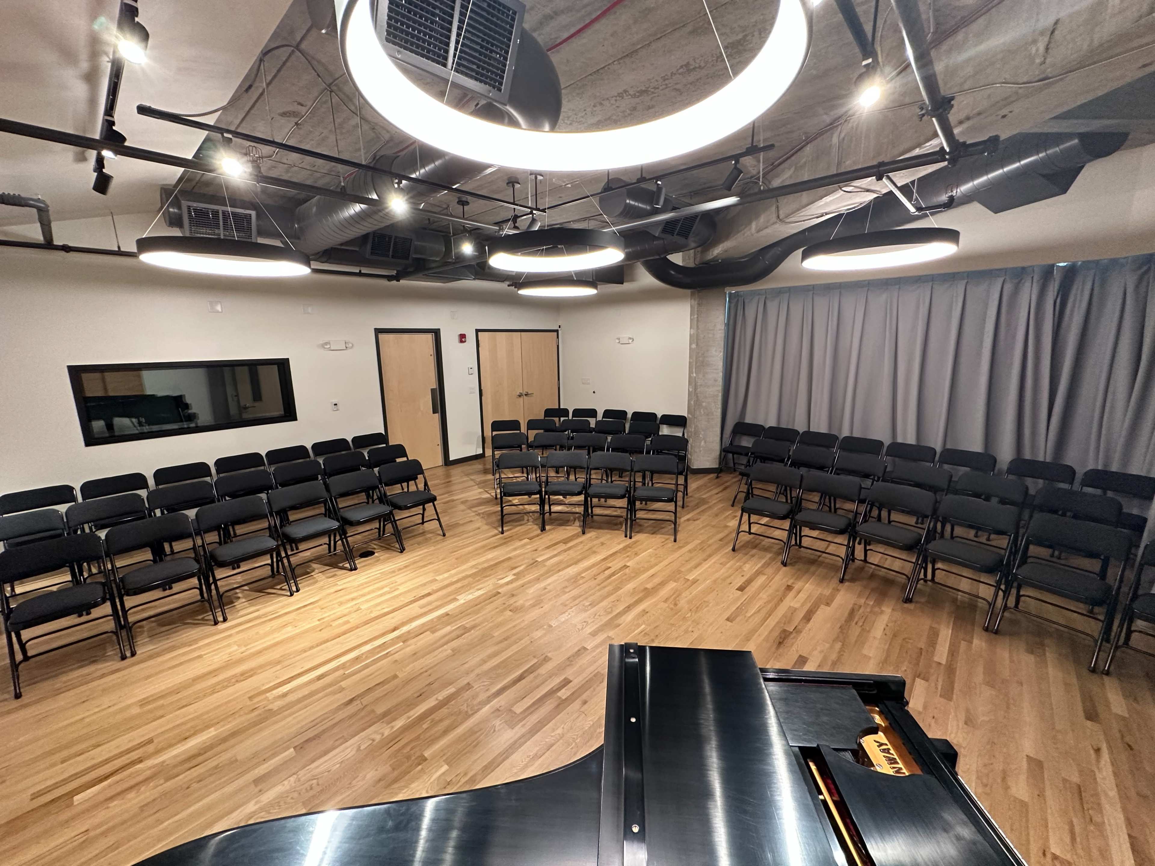 A performance space features rows of black folding chairs arranged in a semicircle around a grand piano, with a curtain and acoustic panels in the background.