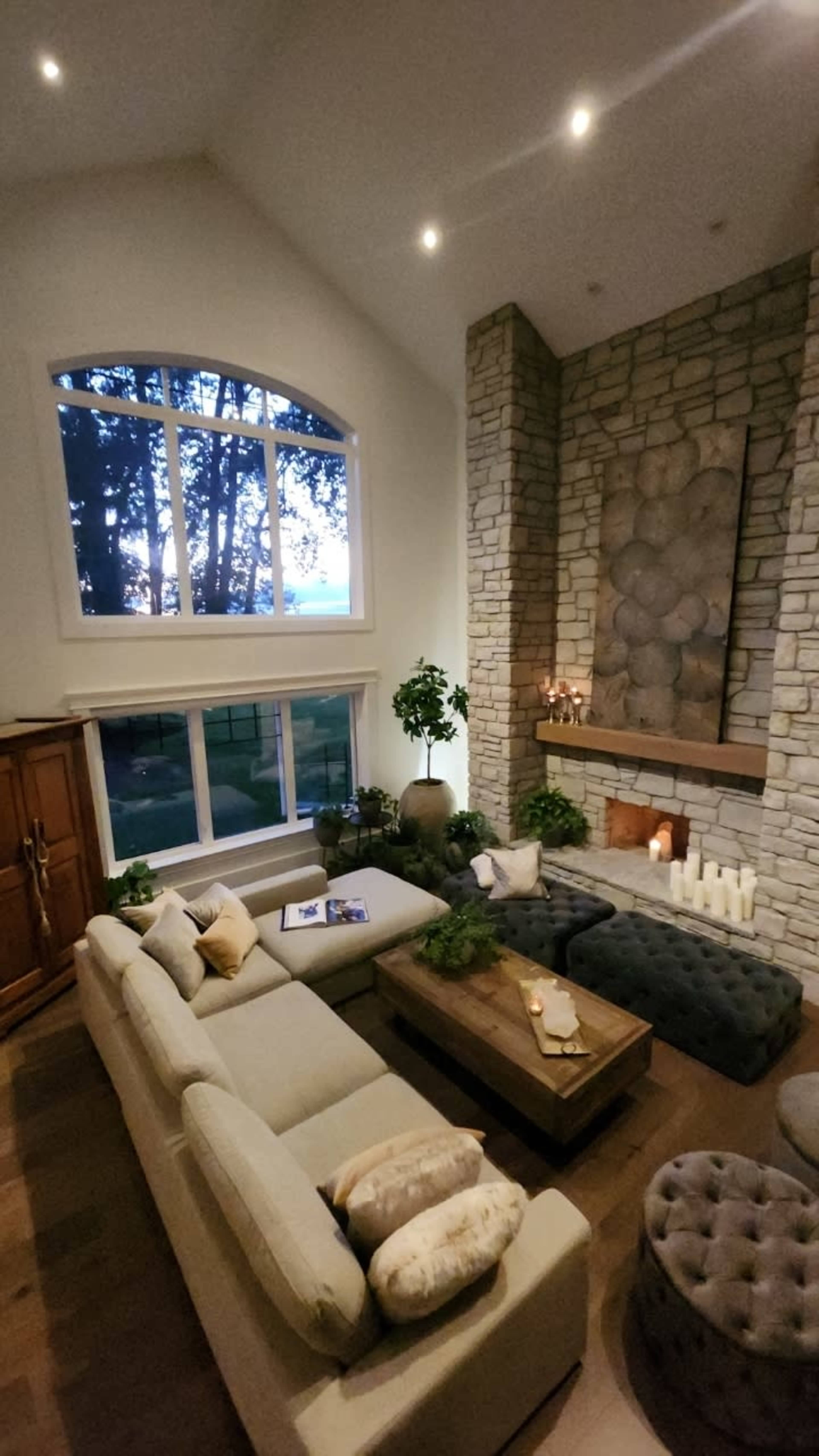 A spacious living room features a large sectional sofa, a stone fireplace, and large windows that provide a view of the outdoors.