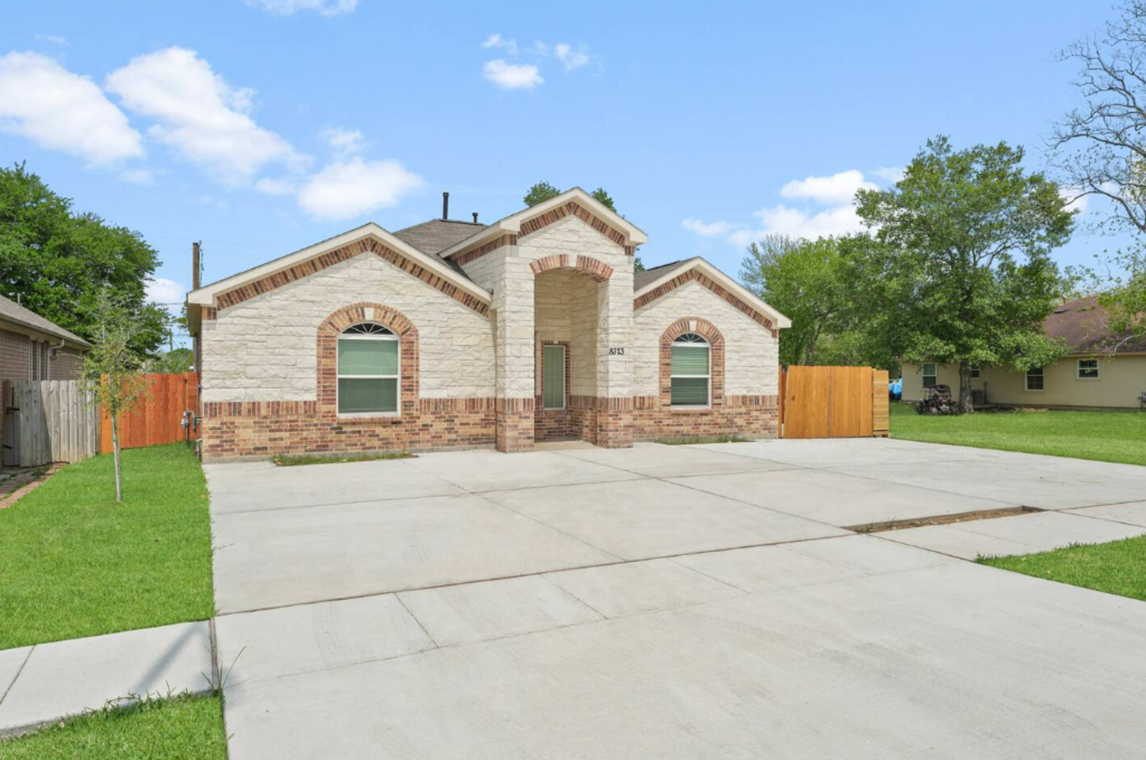 The image shows a single-story house with a brick and stone facade, large front windows, and a spacious concrete driveway.