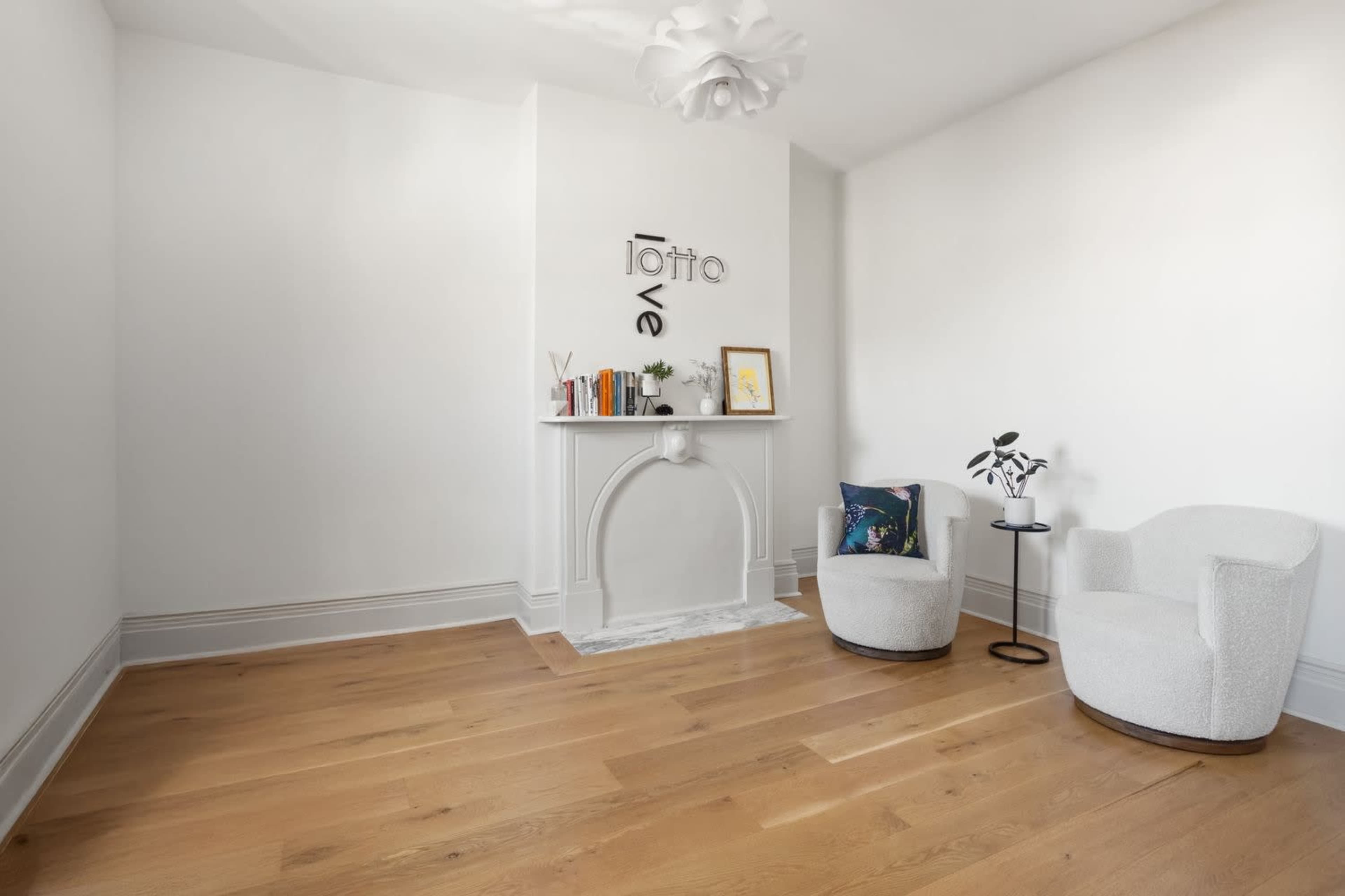The image shows a minimalistic room with a wooden floor, featuring a decorative fireplace, a small shelf with books and a framed picture, and two soft seating chairs.