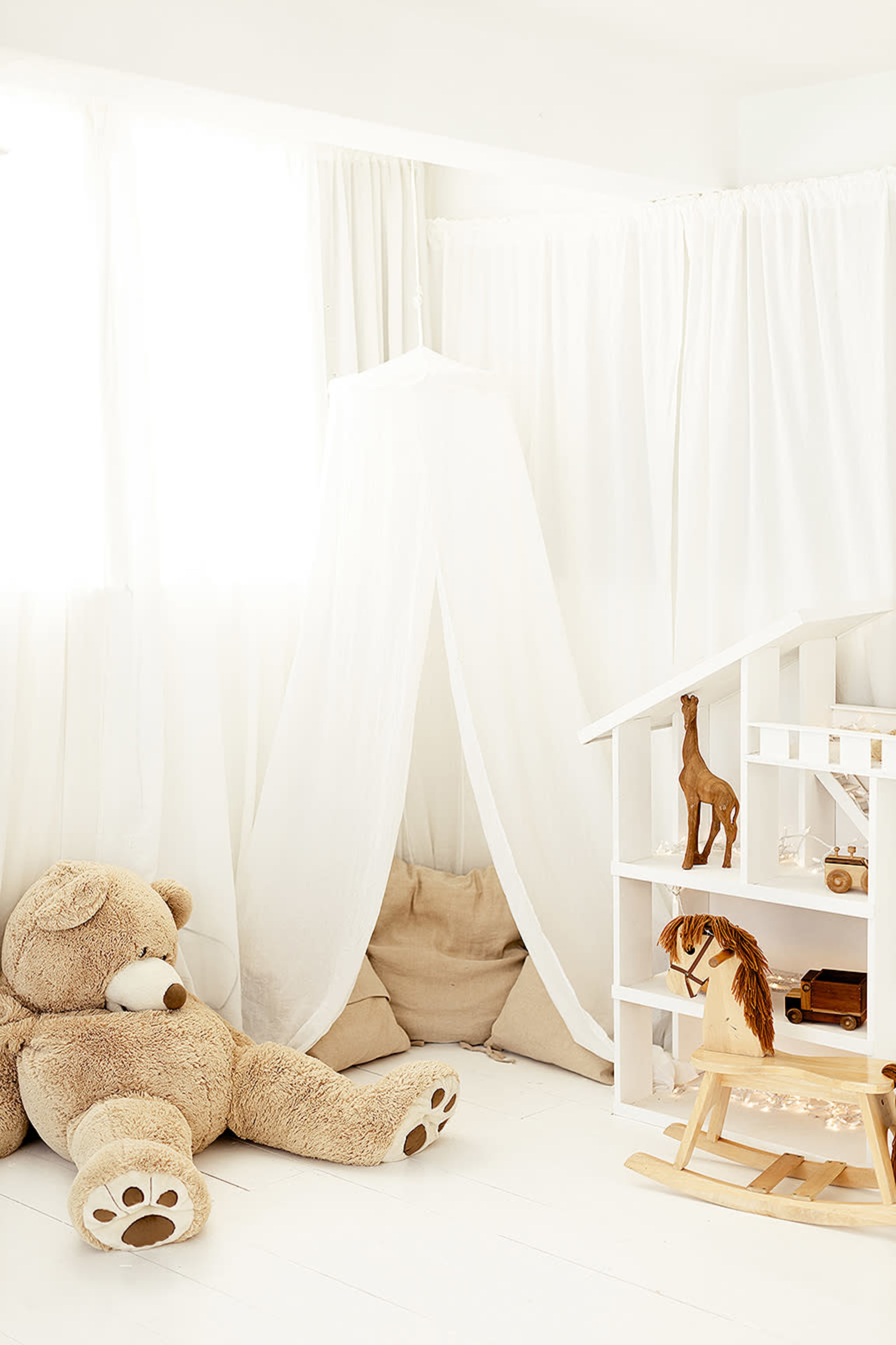 A cozy children's corner features a large teddy bear, a canopy draped in white fabric, and a small wooden rocking horse beside a white staircase.