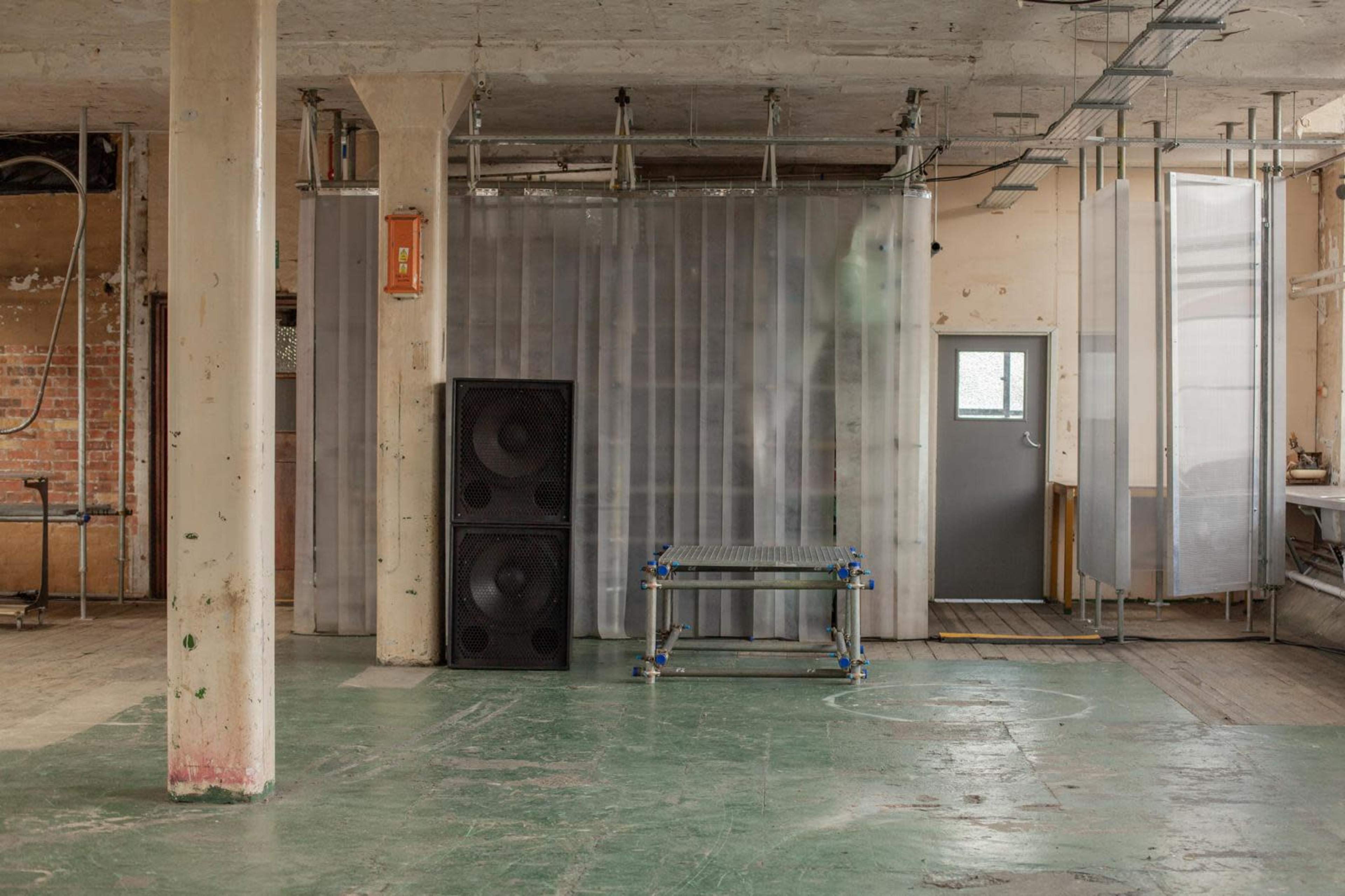 The image shows an industrial interior space with a gray floor, a large speaker stack, and a partition made of transparent material, along with a door and some exposed brickwork.