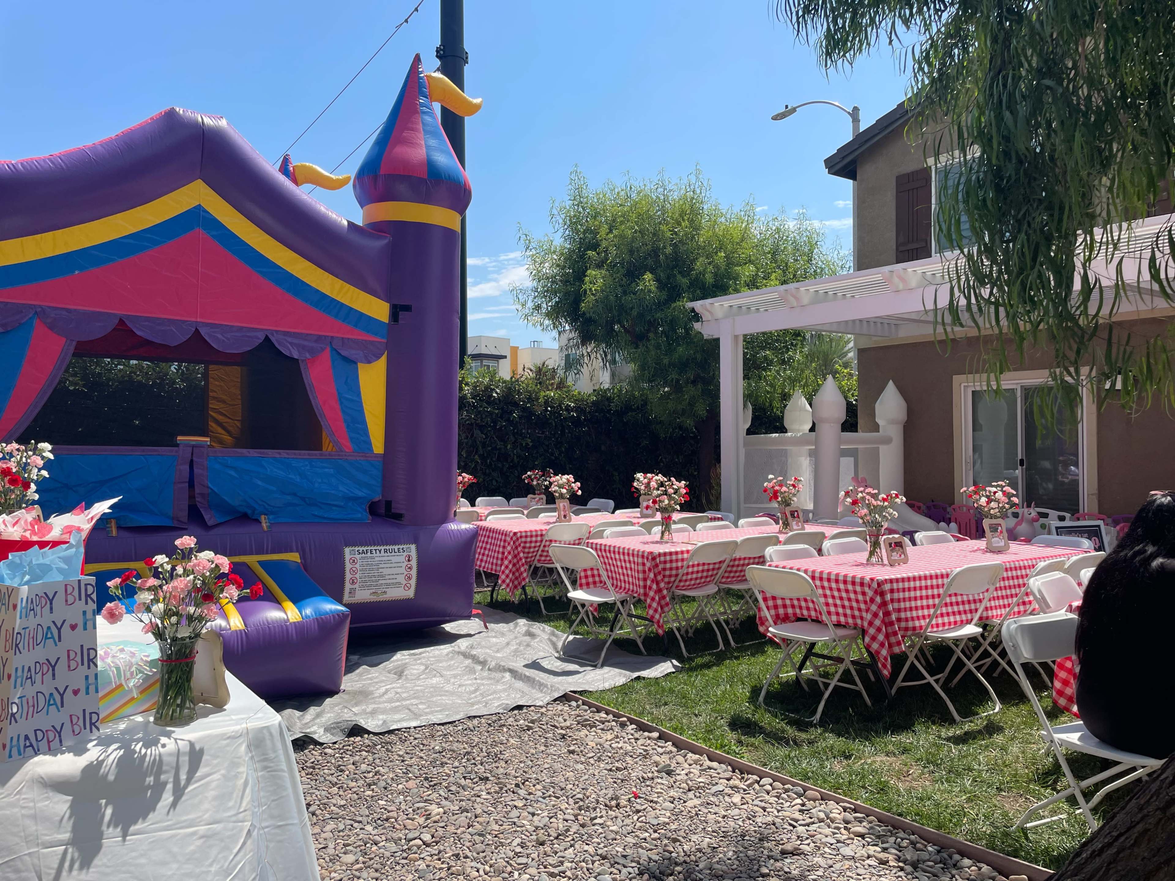 A backyard party setup features a colorful bounce house and tables dressed in checkered tablecloths surrounded by decorations and flowers.
