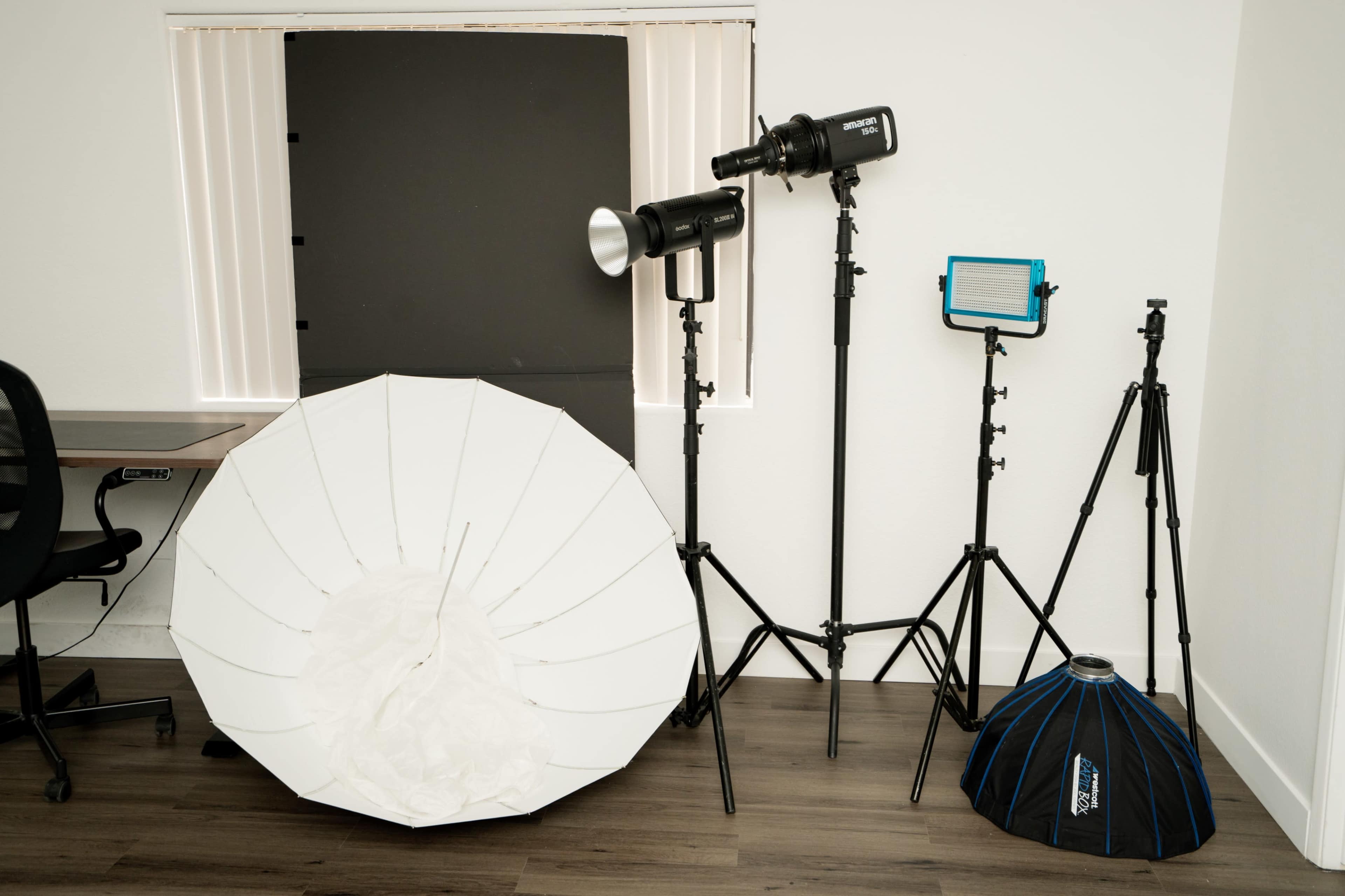 A photography setup with lighting equipment, including an umbrella softbox, a strobe light, and LED panels on tripods, positioned next to a desk in a room.