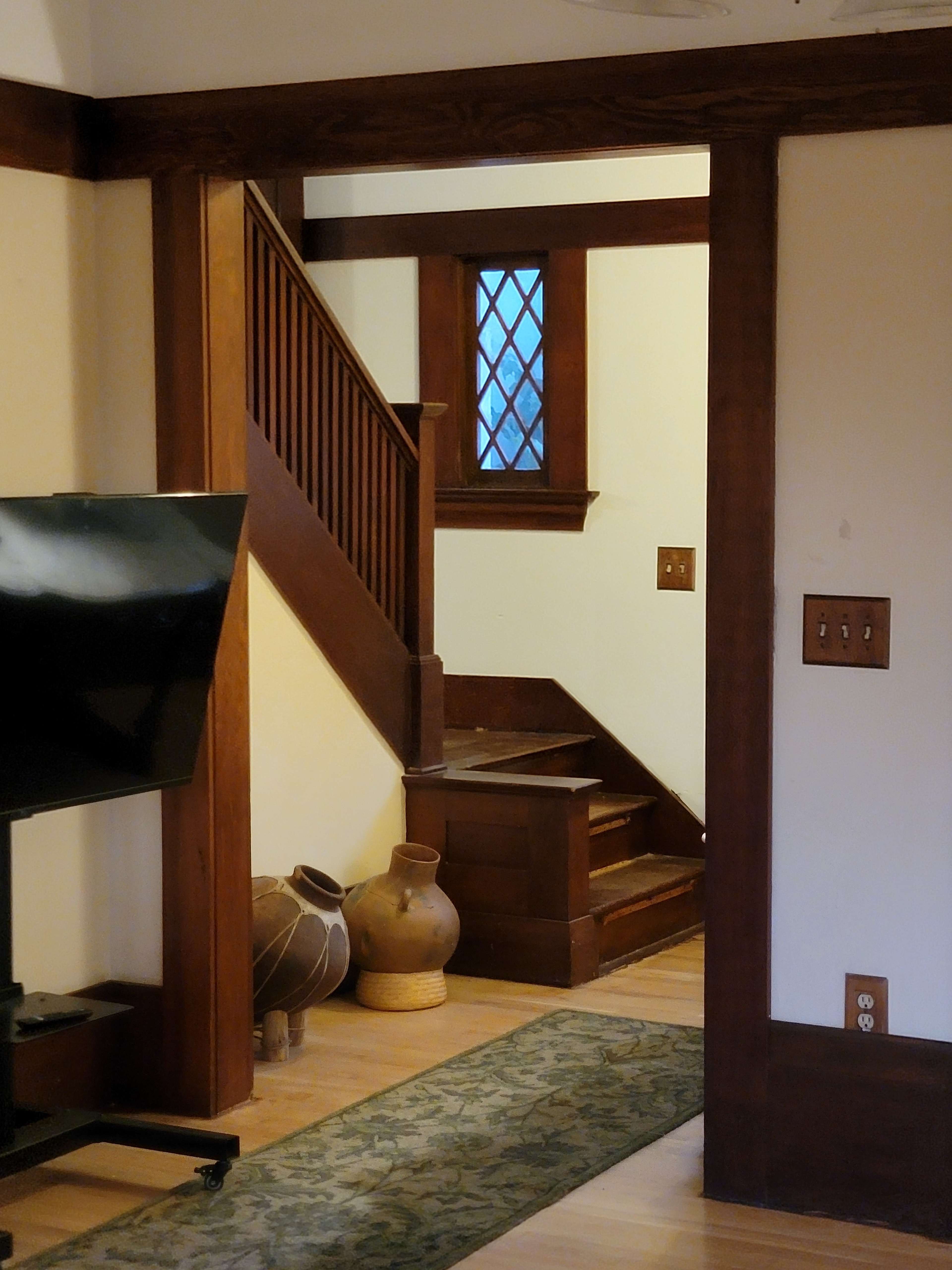 The image shows a hallway with wooden walls, featuring a staircase that leads to a second floor, alongside a television and decorative pottery in the foreground.
