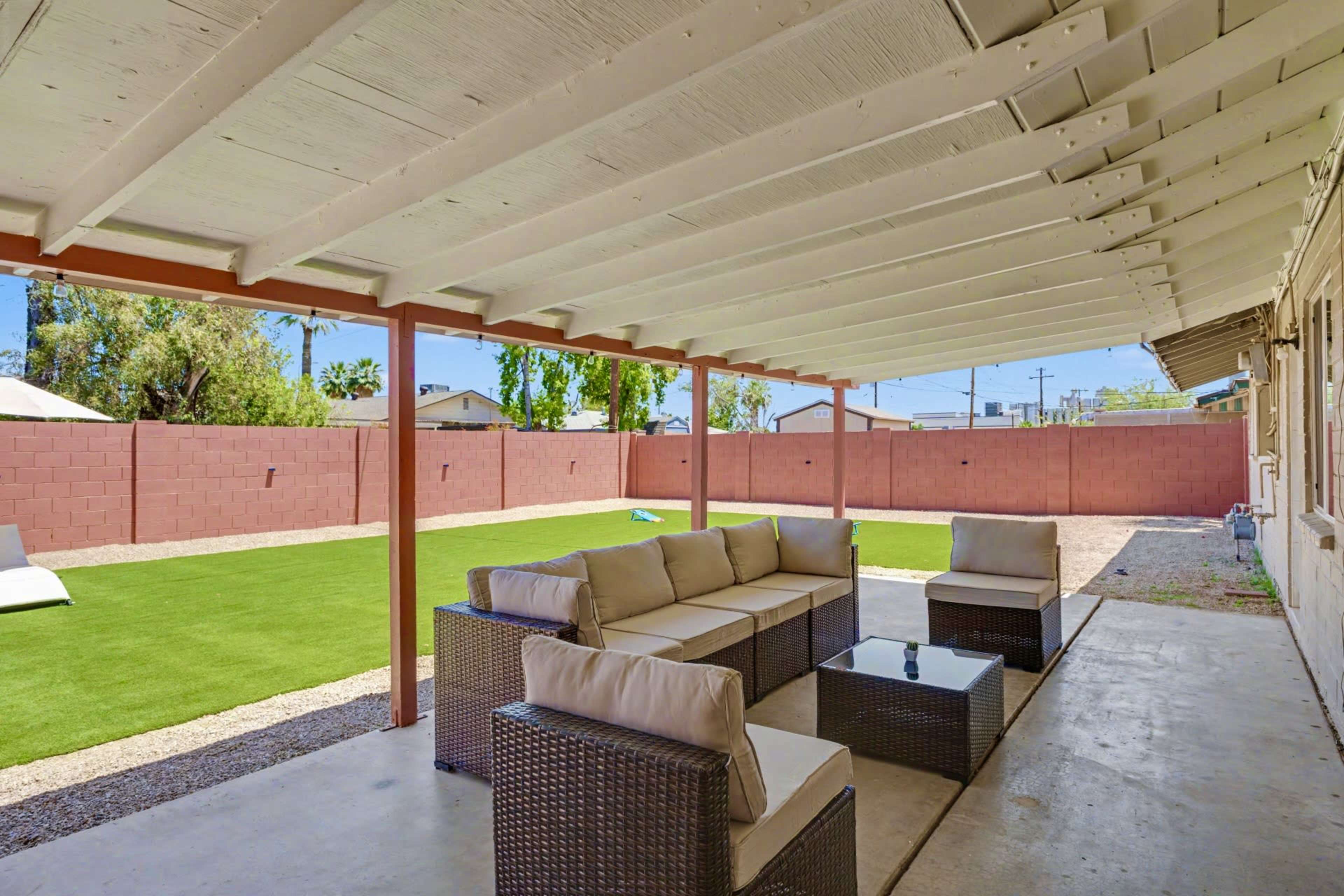 The image shows a covered outdoor patio with a seating area consisting of a sofa and coffee table, overlooking a grassy backyard with a brown wall in the background.