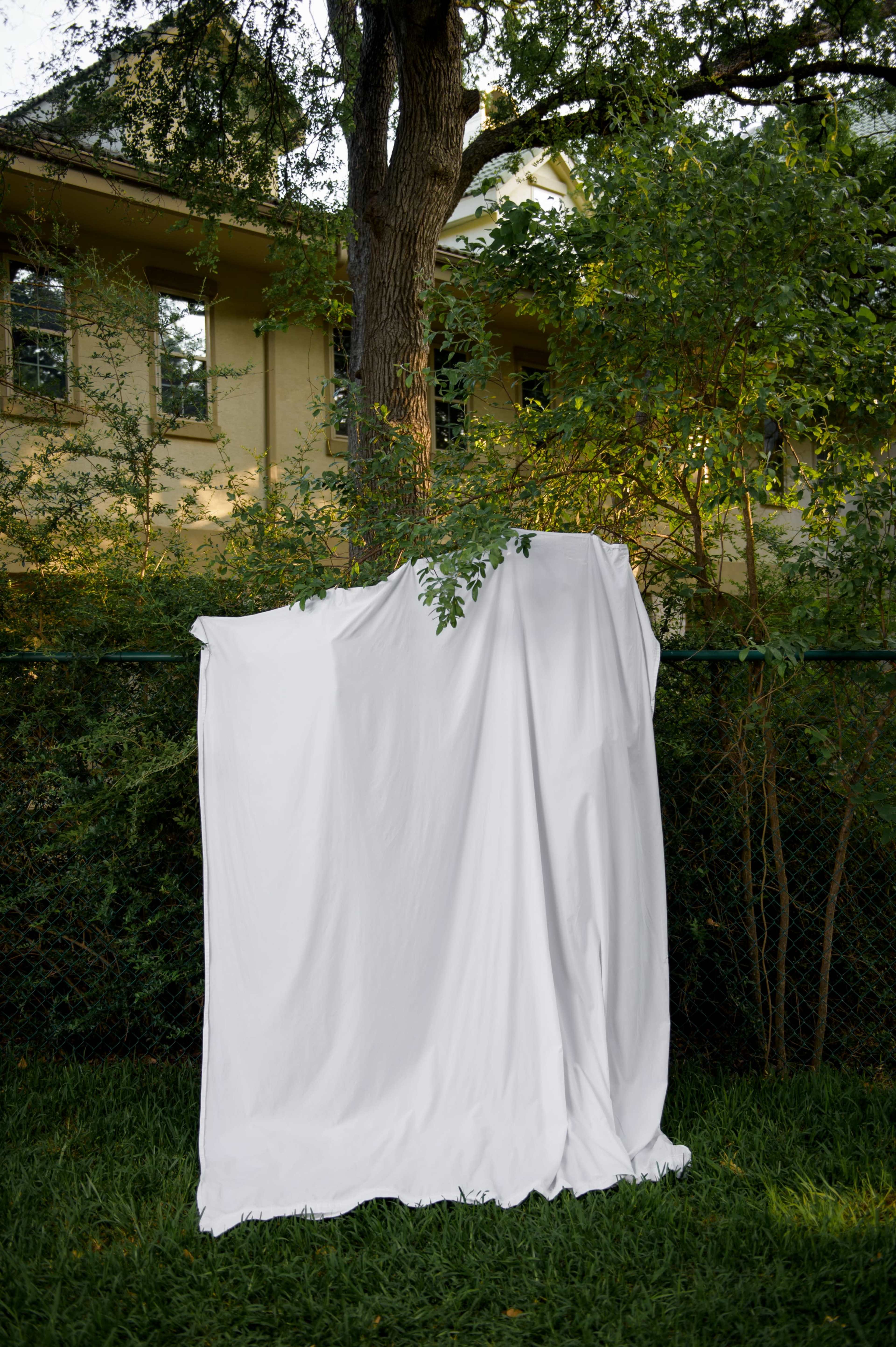 A white sheet is hung on a fence amidst green foliage and a house in the background.