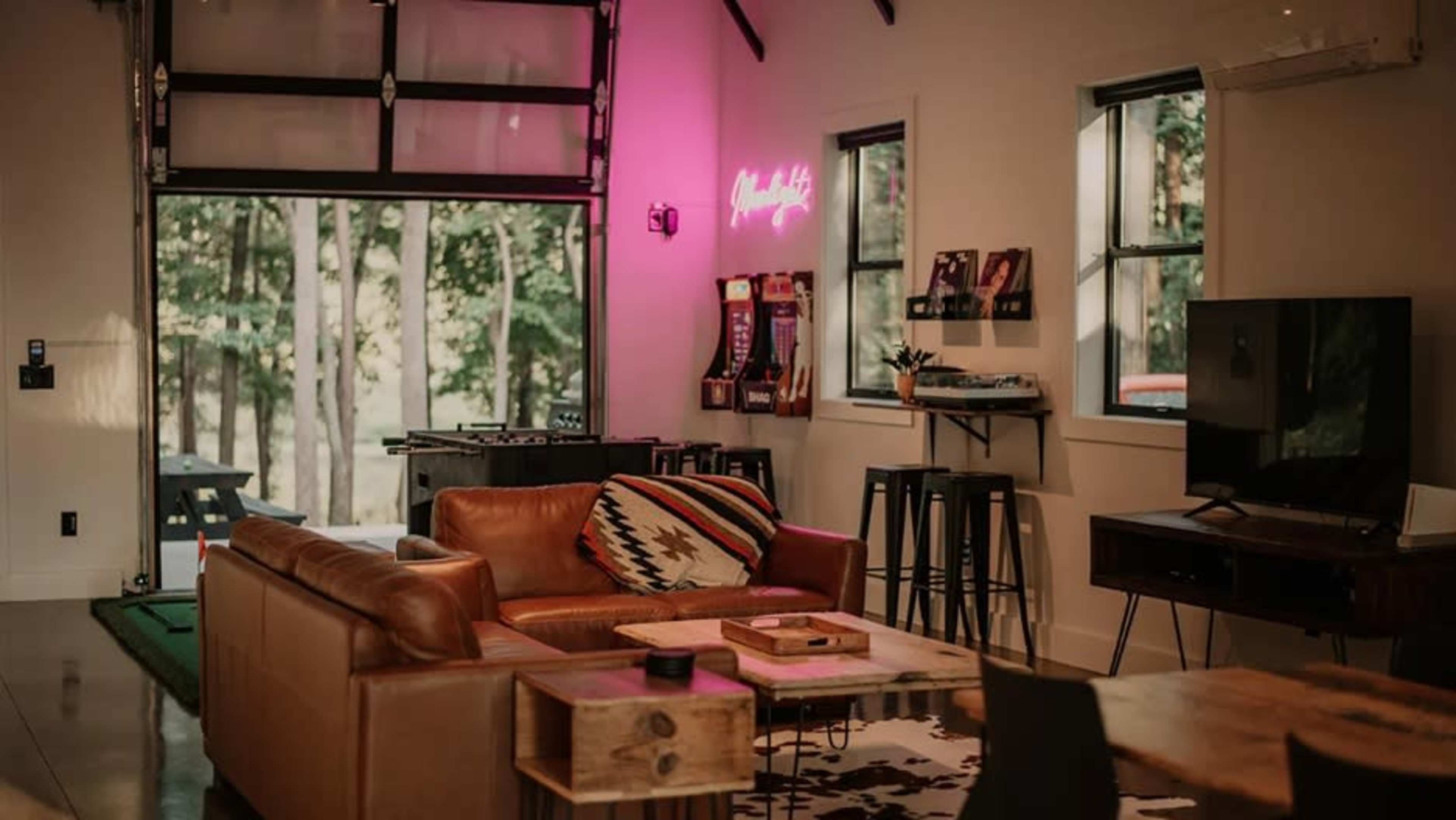 The image shows a modern living room with a leather sofa, a wooden coffee table, bar stools, and a large window revealing trees outside.
