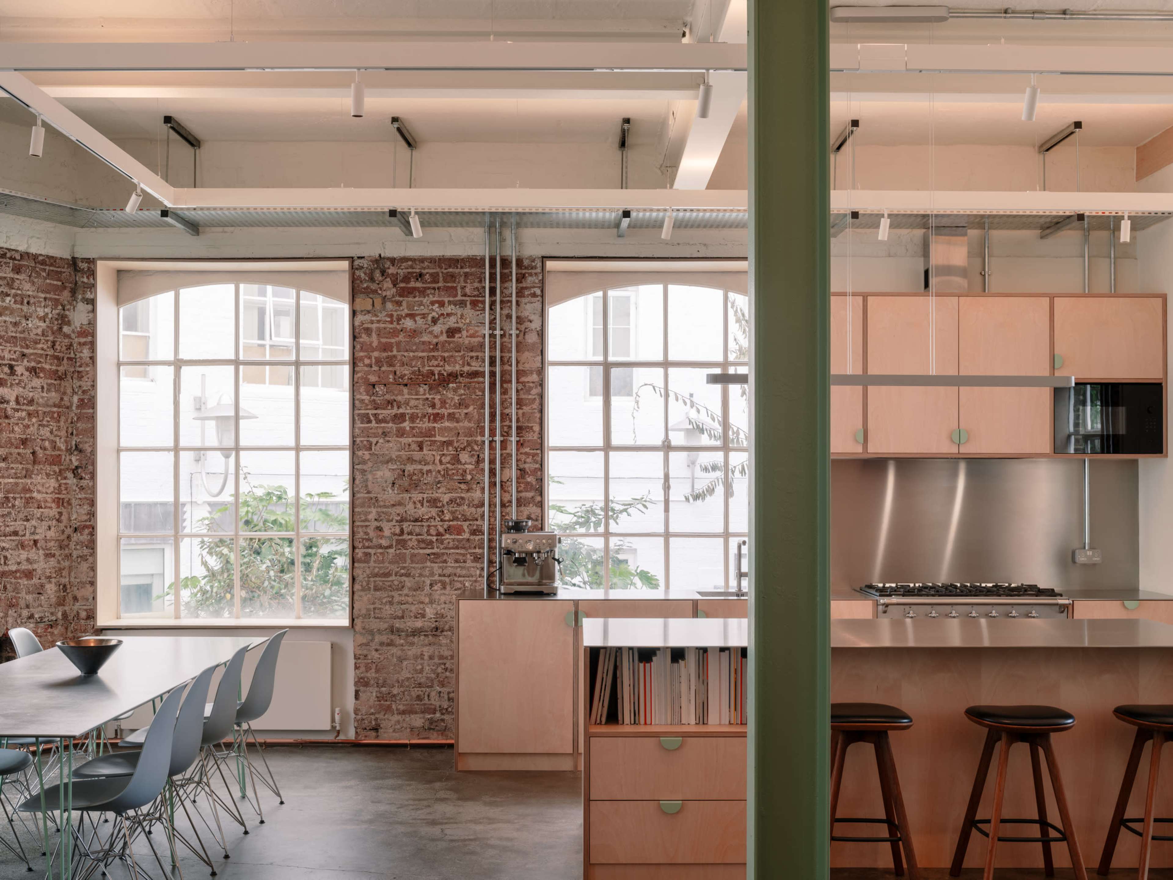 The image shows a modern kitchen and dining area with a combination of wooden cabinetry, a stainless steel stove, and large windows showcasing a brick wall backdrop.