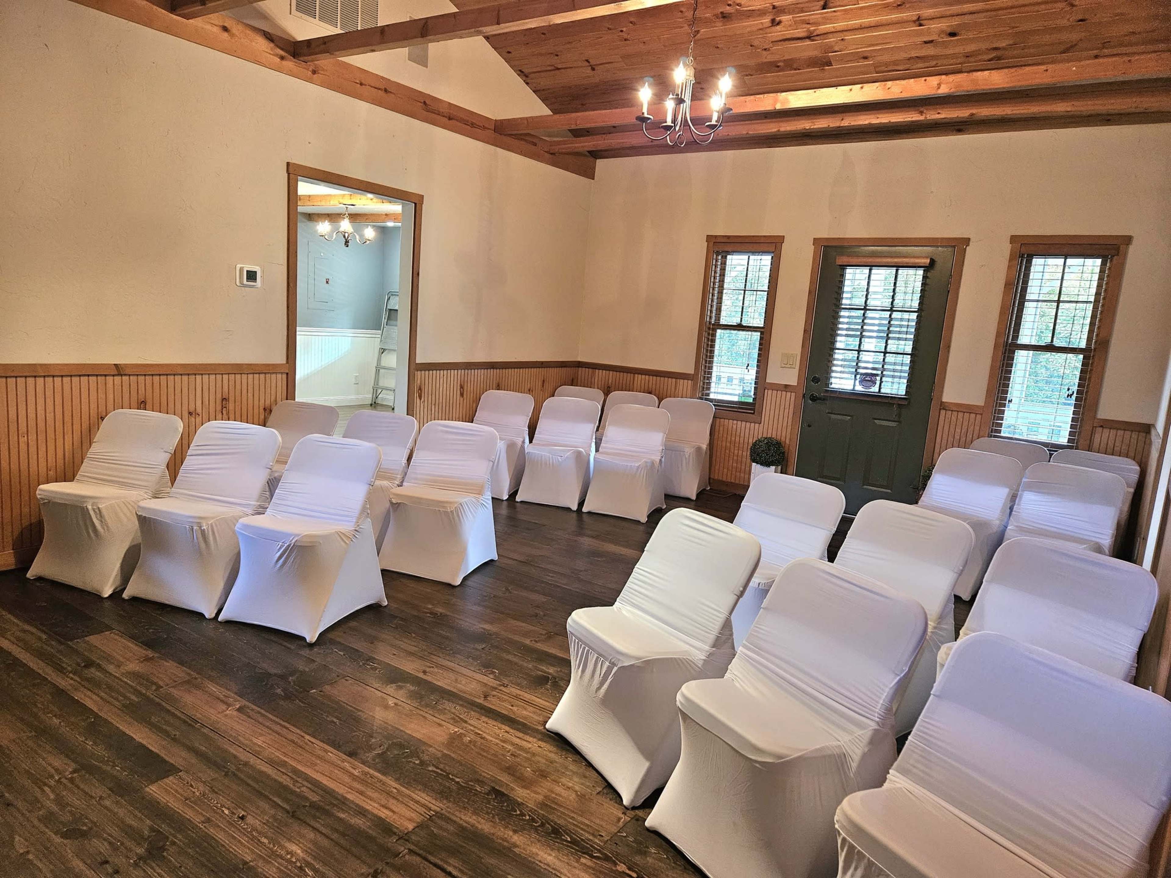 The image shows a room with wooden beams and a dark wooden floor, set up with rows of white-seated chairs facing a door and windows.