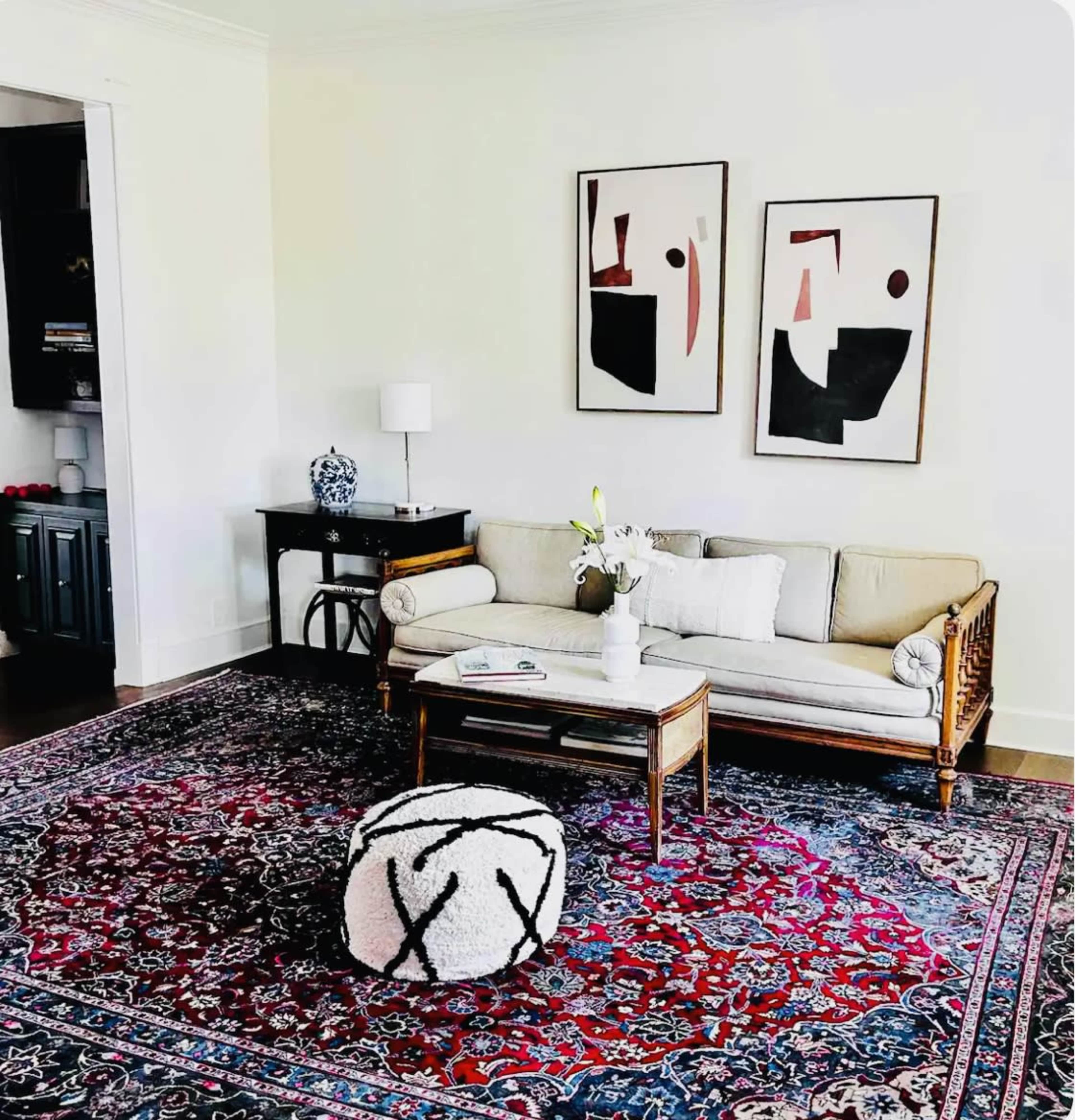 A living room features a beige sofa, a wooden coffee table, a white lamp, and two abstract paintings on the wall, all set on a large, patterned area rug.