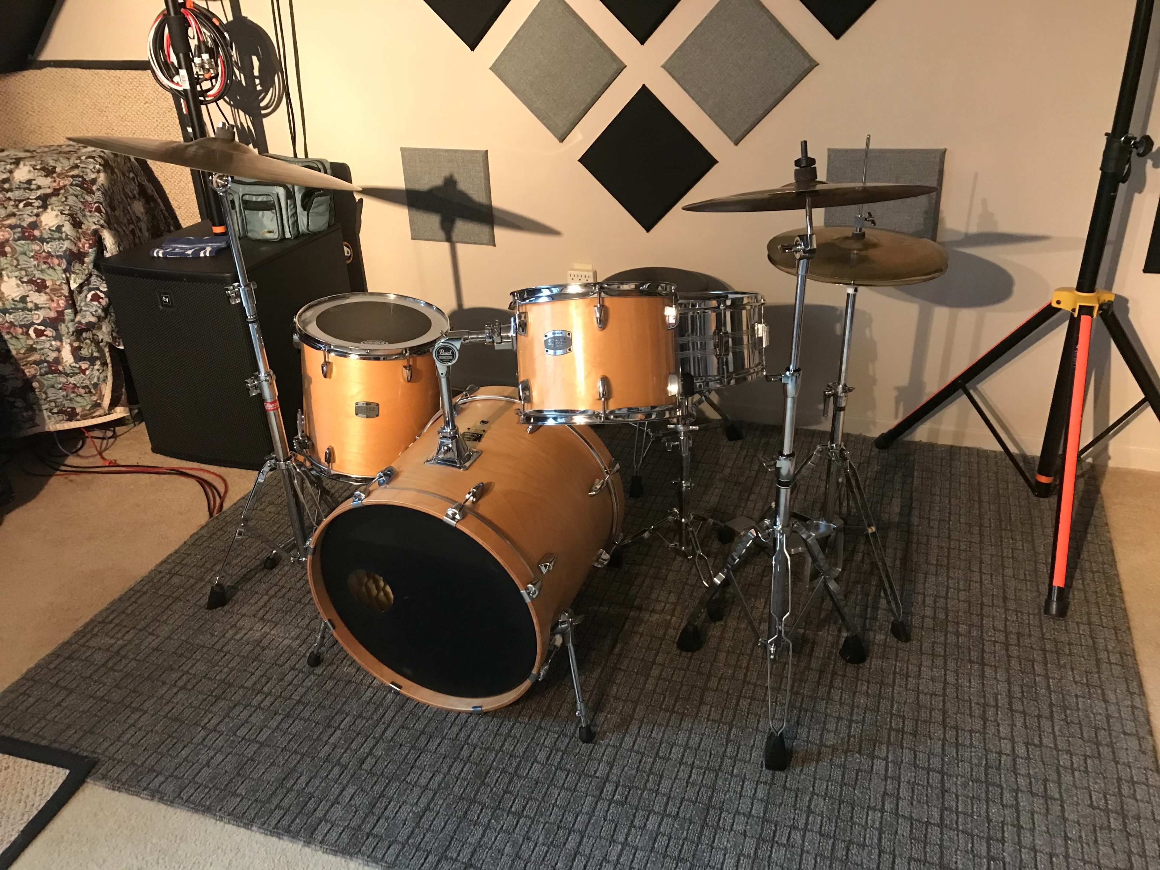 The image shows a wooden drum set with a bass drum, snare drum, and cymbals, positioned on a gray area rug in a music room.
