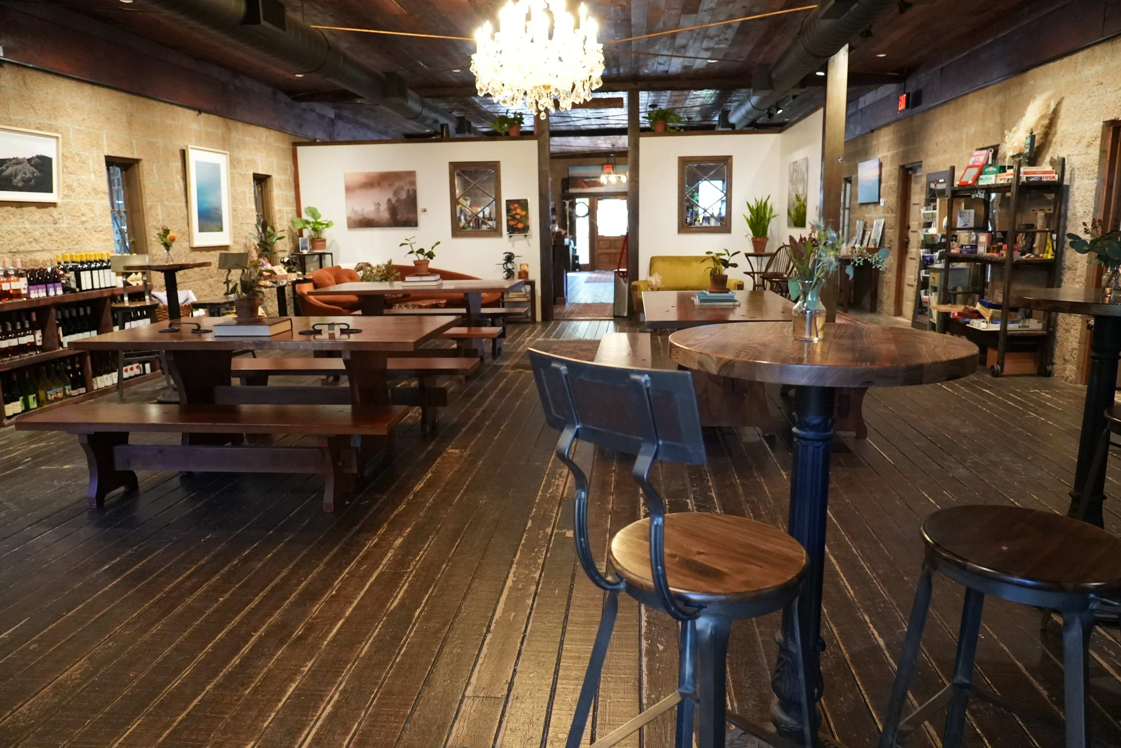 The image shows a spacious interior of a rustic cafe with wooden furniture, tables, and a chandelier, featuring artwork on the walls and plants throughout the room.
