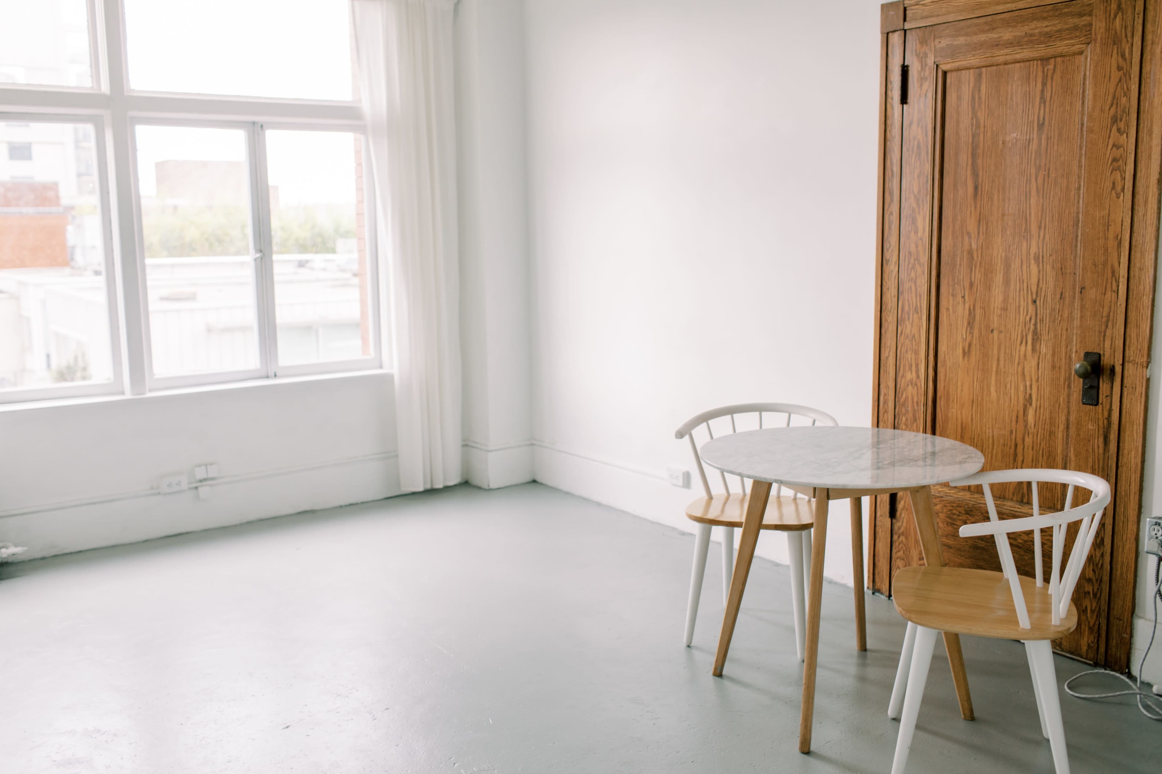 A small, minimalist room features a round marble table with two wooden chairs beside a wooden closet and large windows letting in natural light.