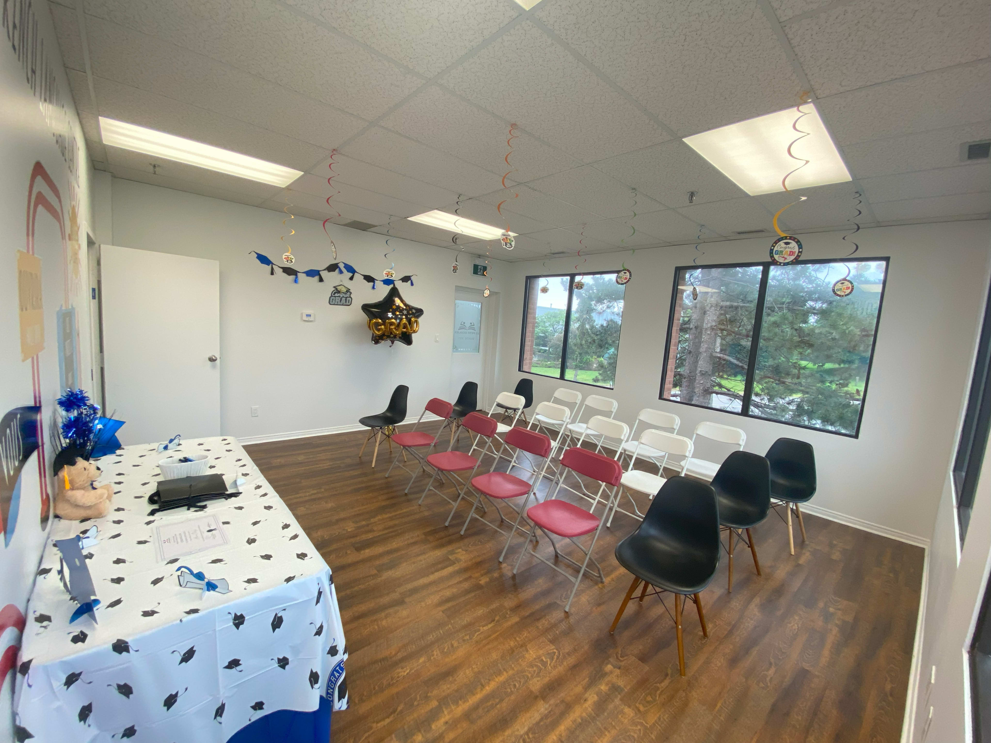 A decorated room with rows of chairs set up for a gathering, featuring a table with a festive tablecloth and graduation-themed decor hanging from the ceiling.
