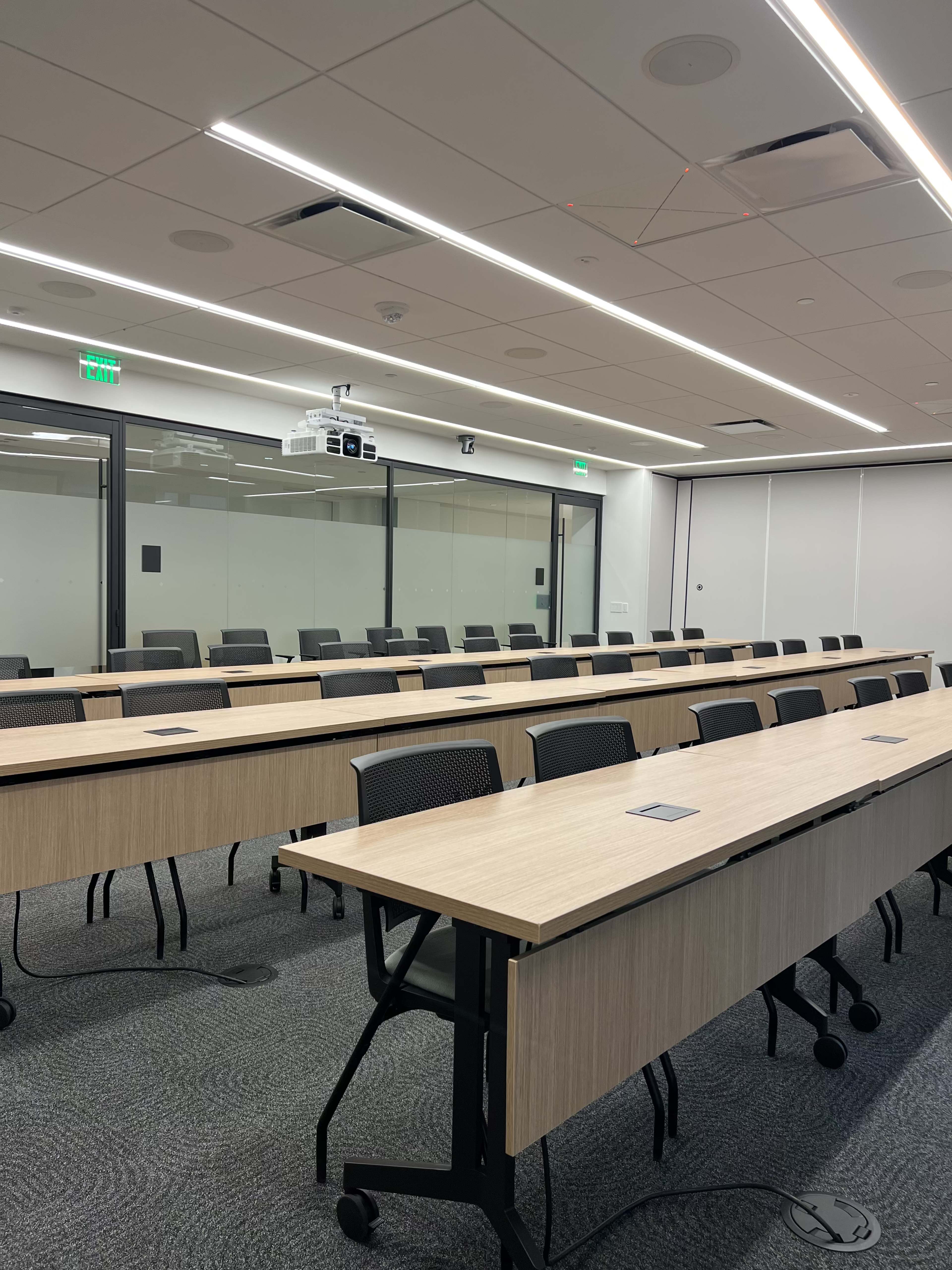 The image shows a modern conference room with rows of tables and black chairs, and a projector mounted on the ceiling.