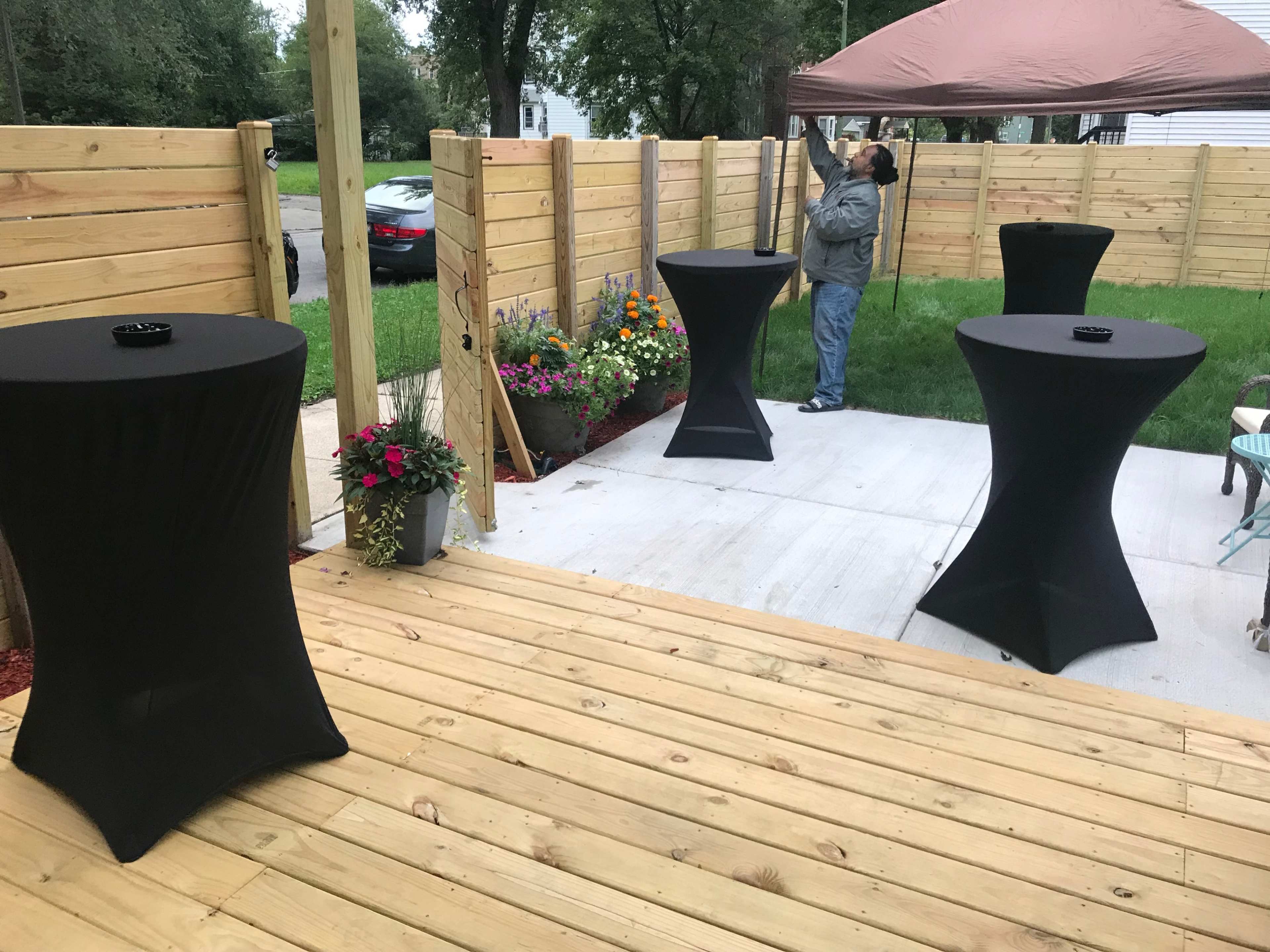 A patio area with three black cocktail tables and a man adjusting a tent in the background, surrounded by wooden fencing and flower beds.