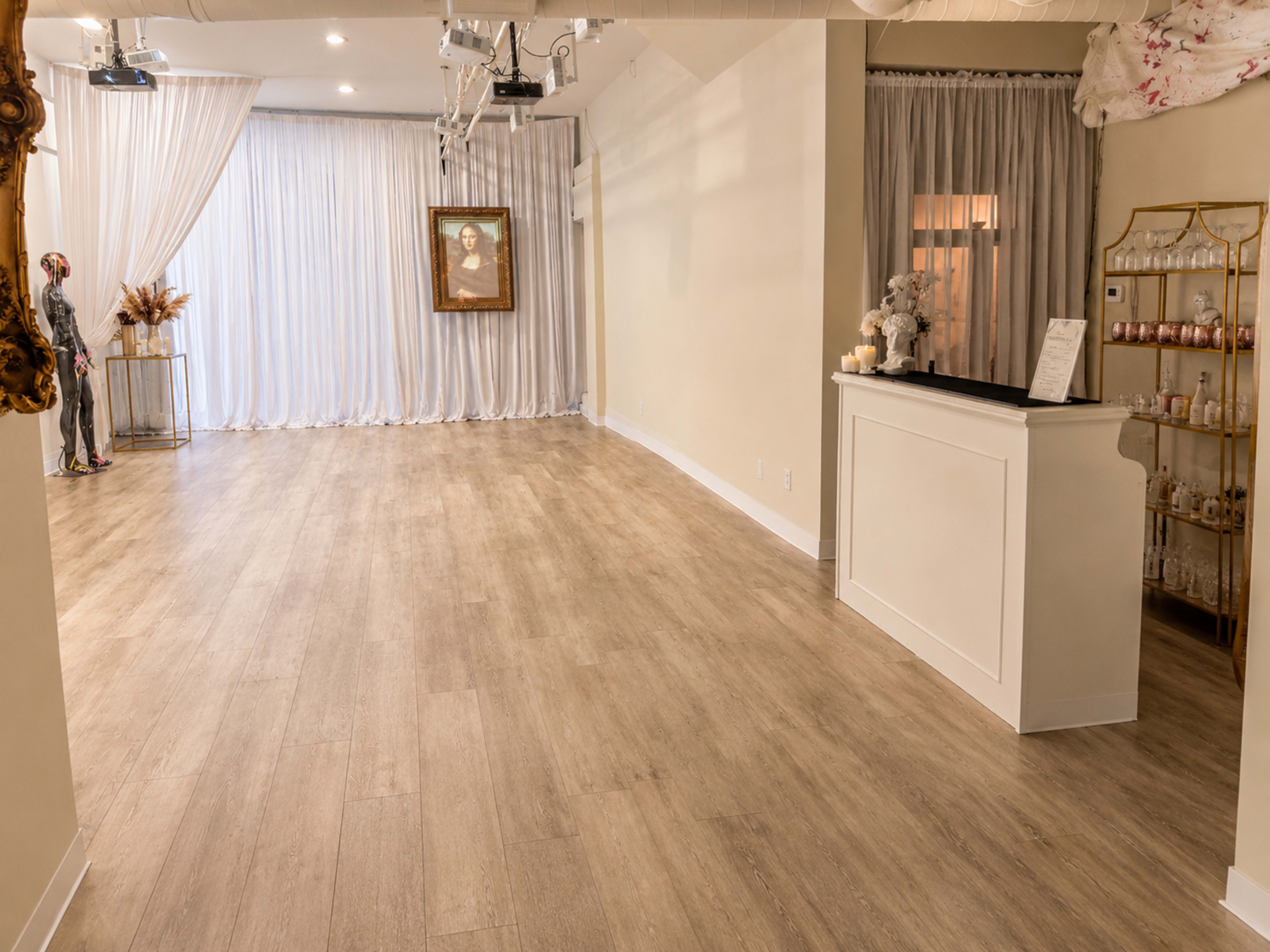 The image shows a spacious, well-lit room with wooden flooring, soft curtains, and a reception counter, along with a decorative frame on the wall.