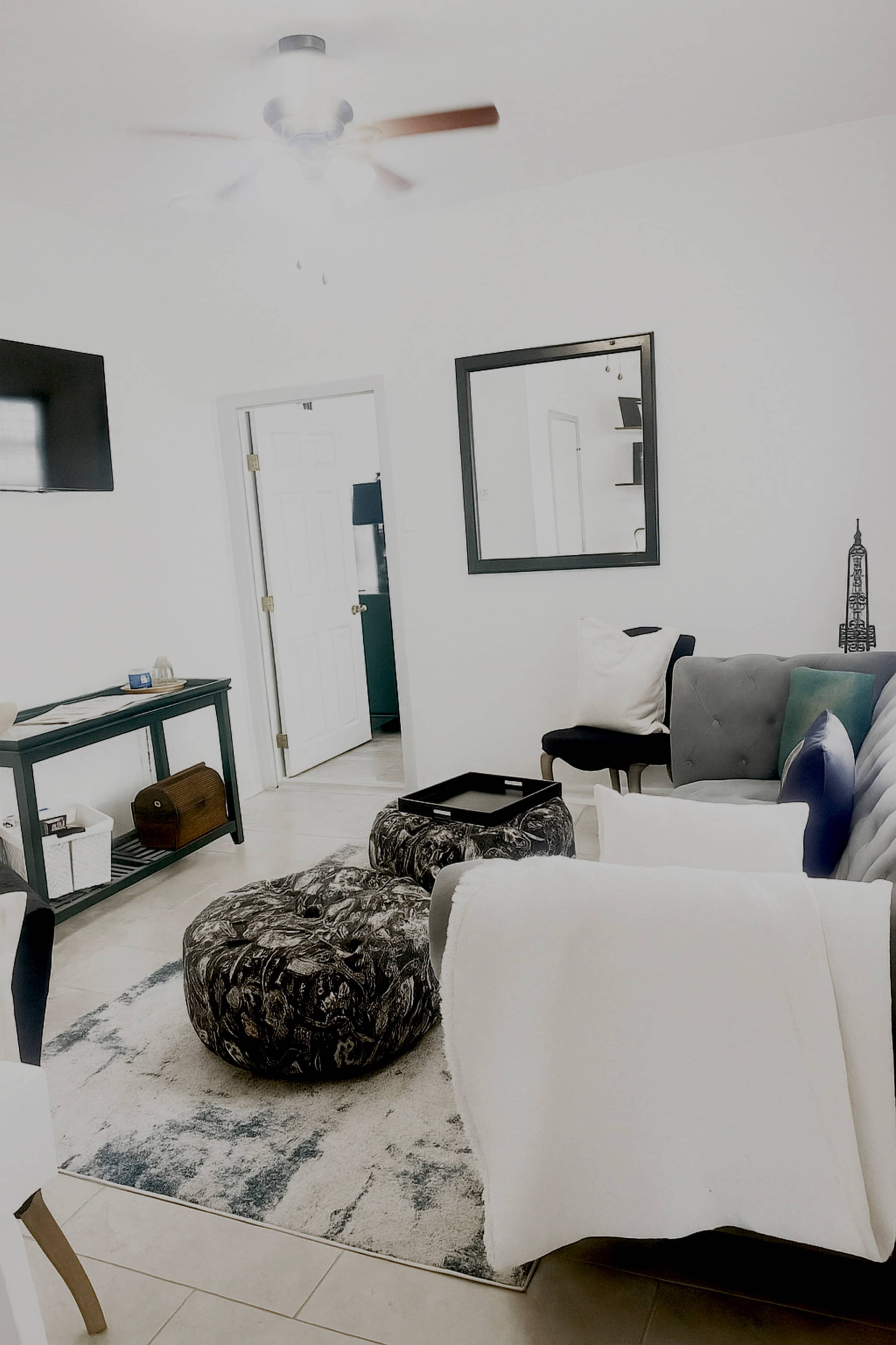 The image shows a clean living room with a green console table, two round ottomans, a gray sofa, and a large mirror on the wall.