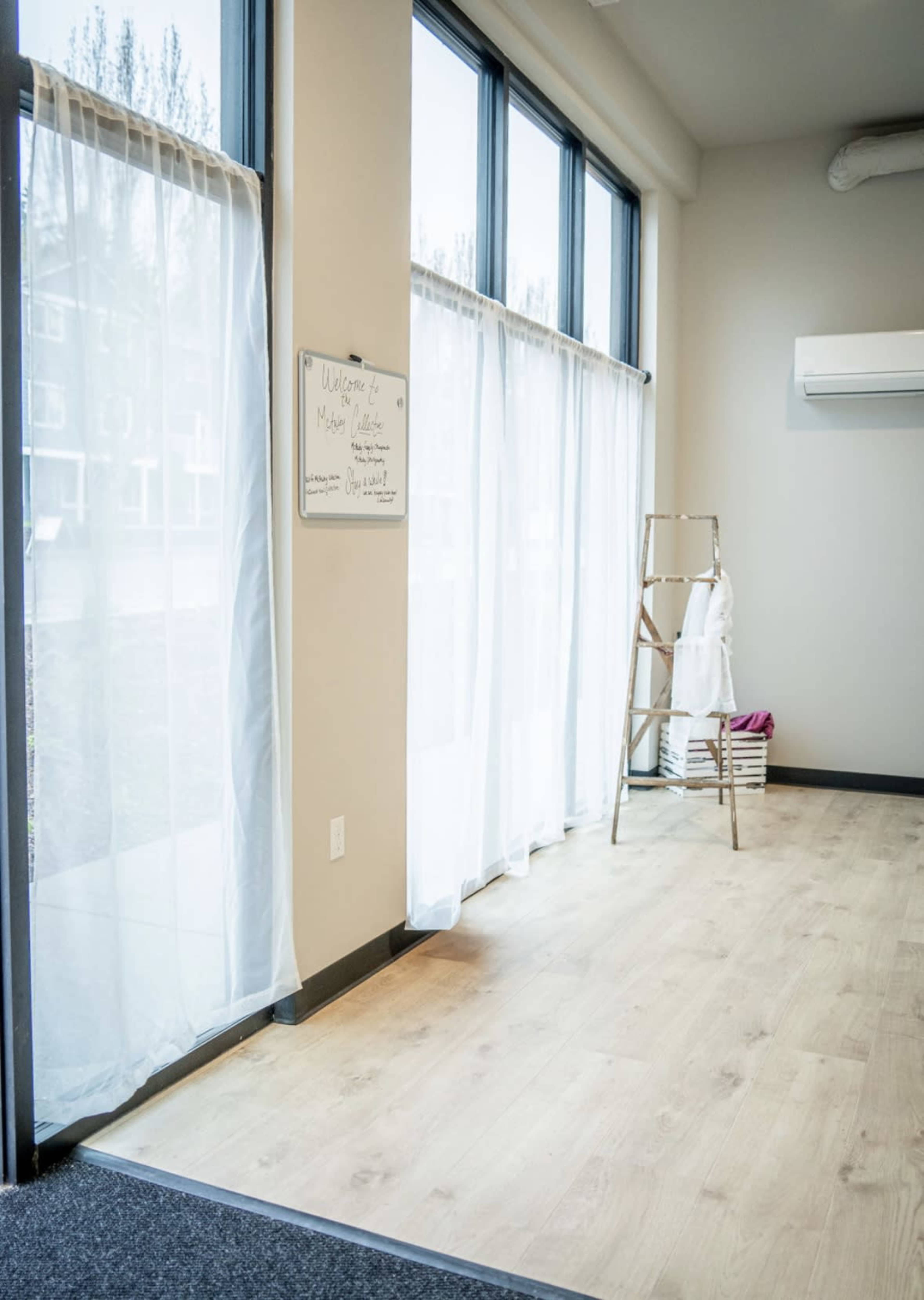 The image shows a spacious room with large windows covered by sheer white curtains, a ladder against the wall, and a sign on a board.