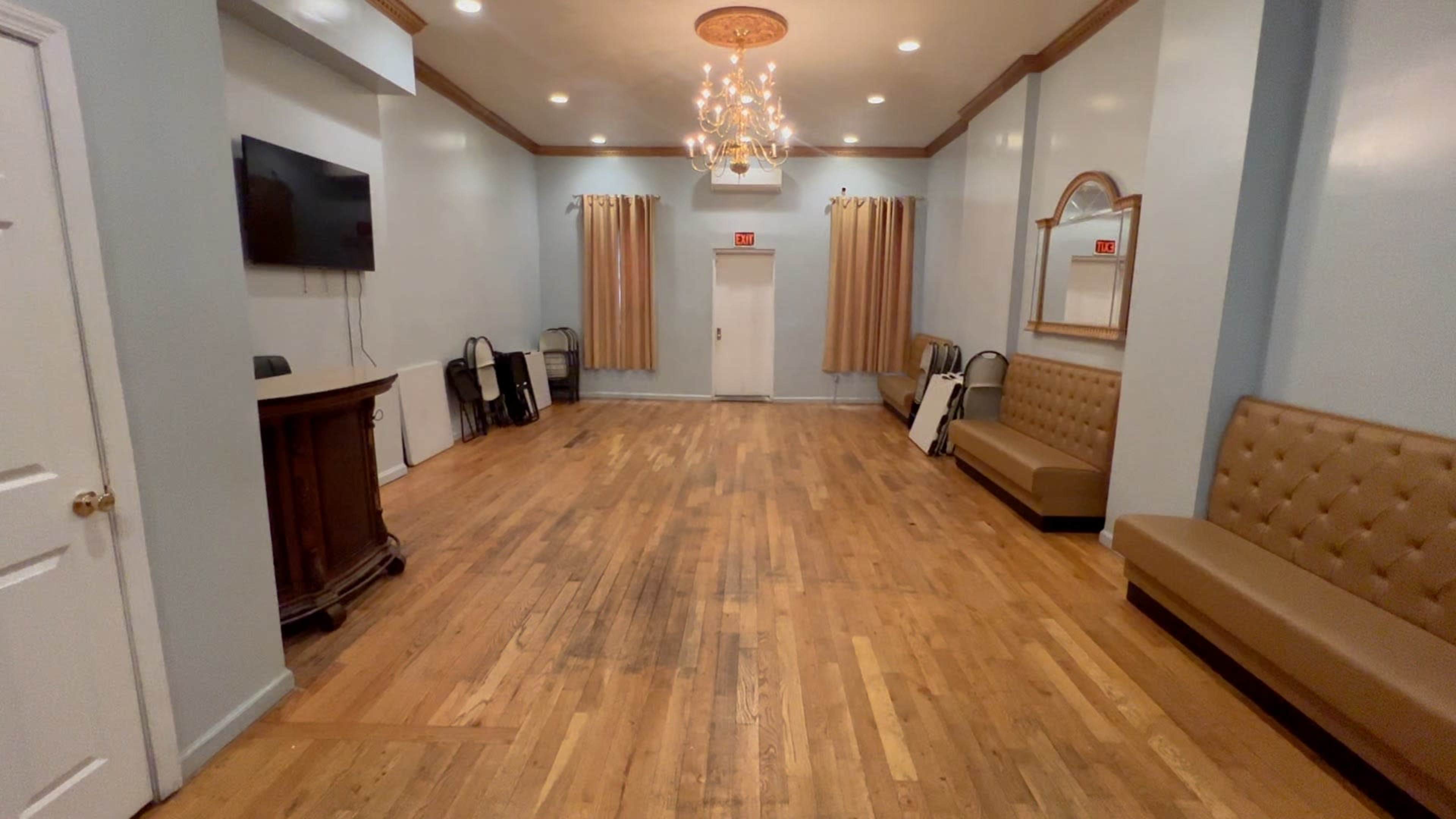 An empty room with wooden flooring, a chandelier, and decorative walls, featuring a bar area and seating along the sides.
