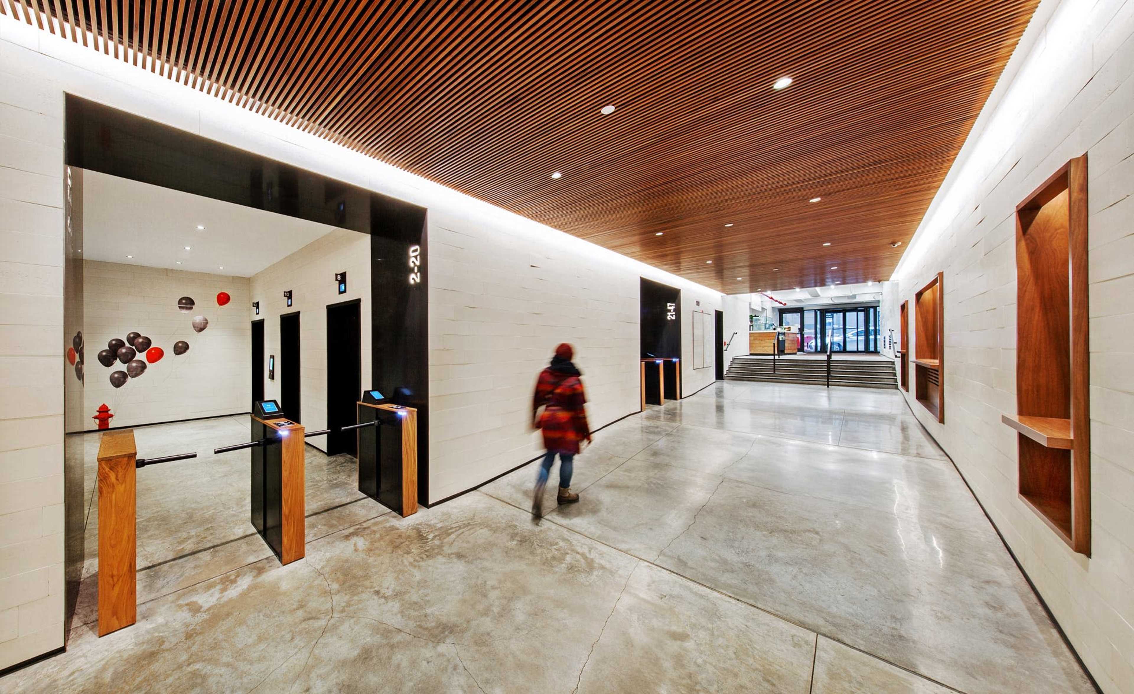 A person walks through a modern hallway featuring elevator doors, wooden accents, and a bright entrance in the background.