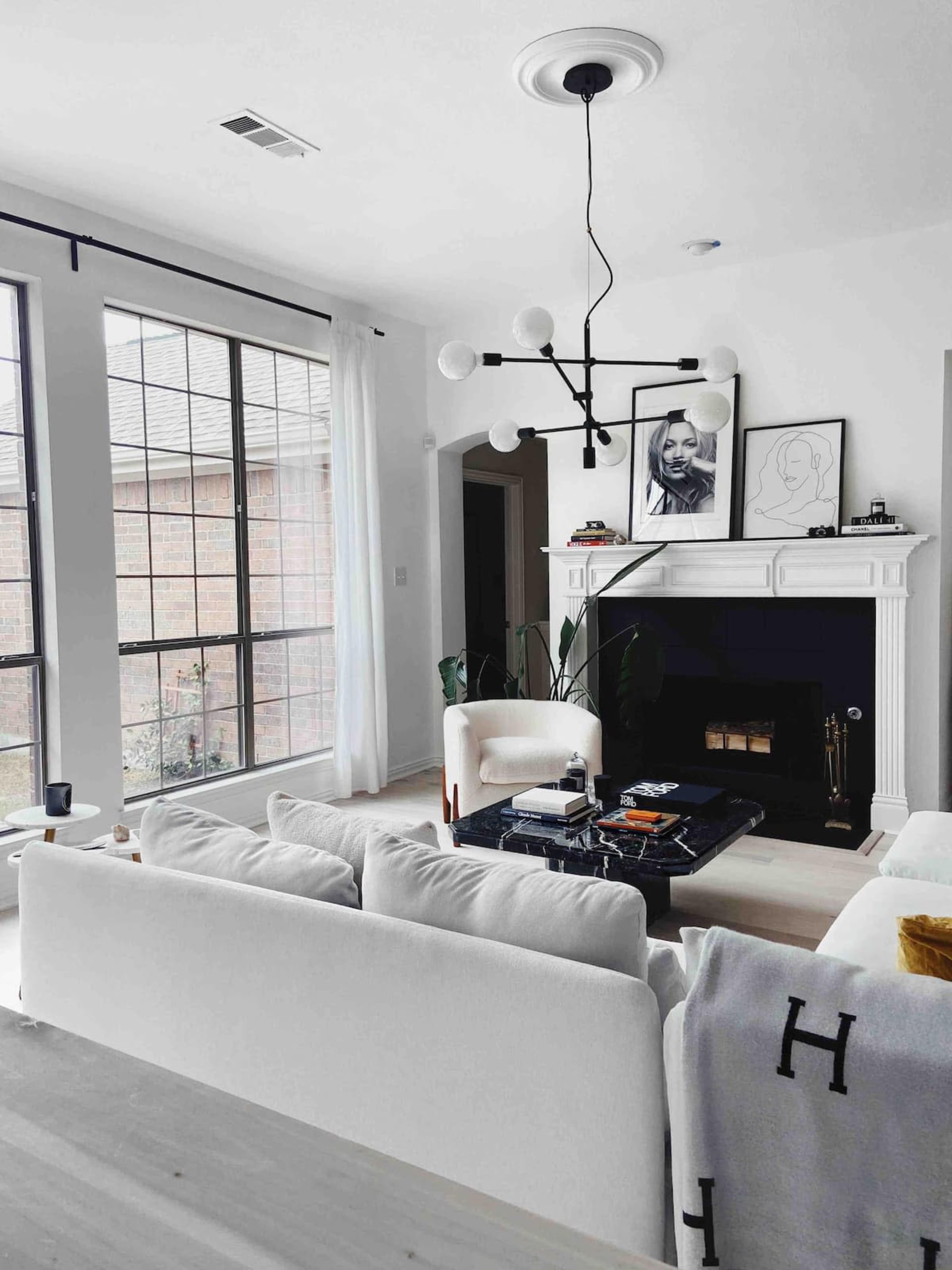 The image shows a modern living room with large windows, a white sofa, a black coffee table, and decorative items, including a framed artwork on the wall above a fireplace.