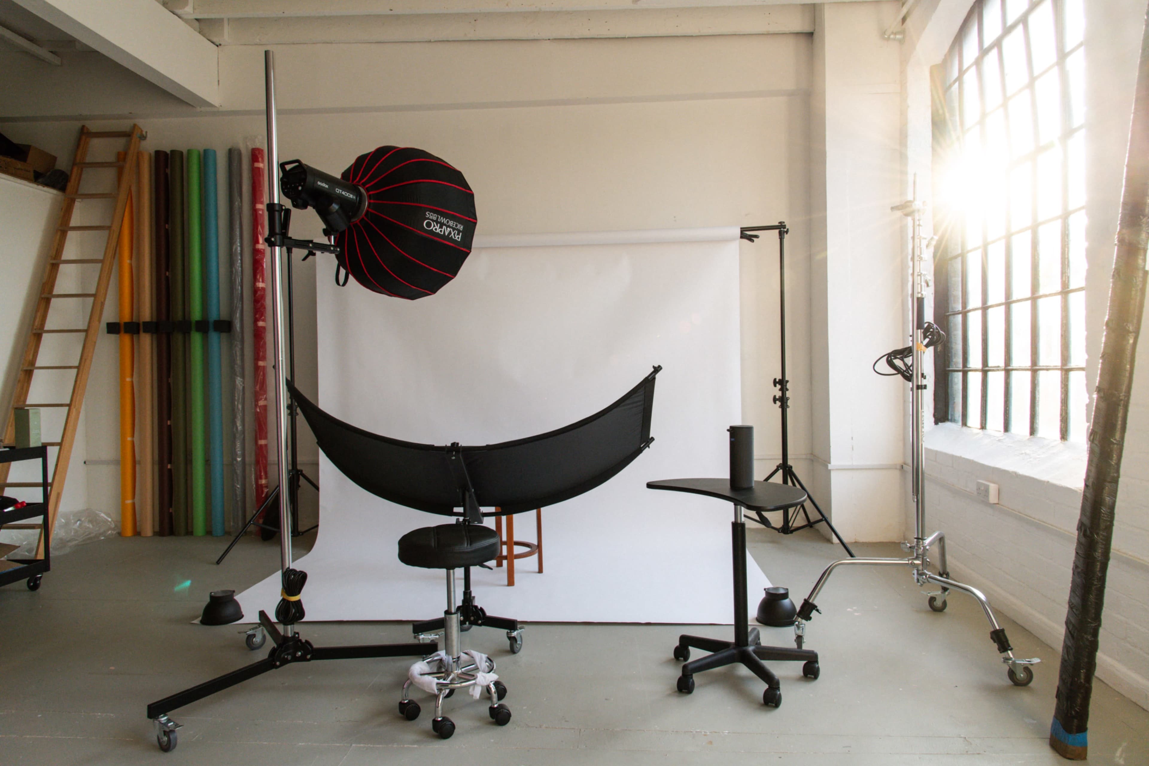 A photography studio with a backdrop, lighting equipment, and various chairs arranged in front of a large window.