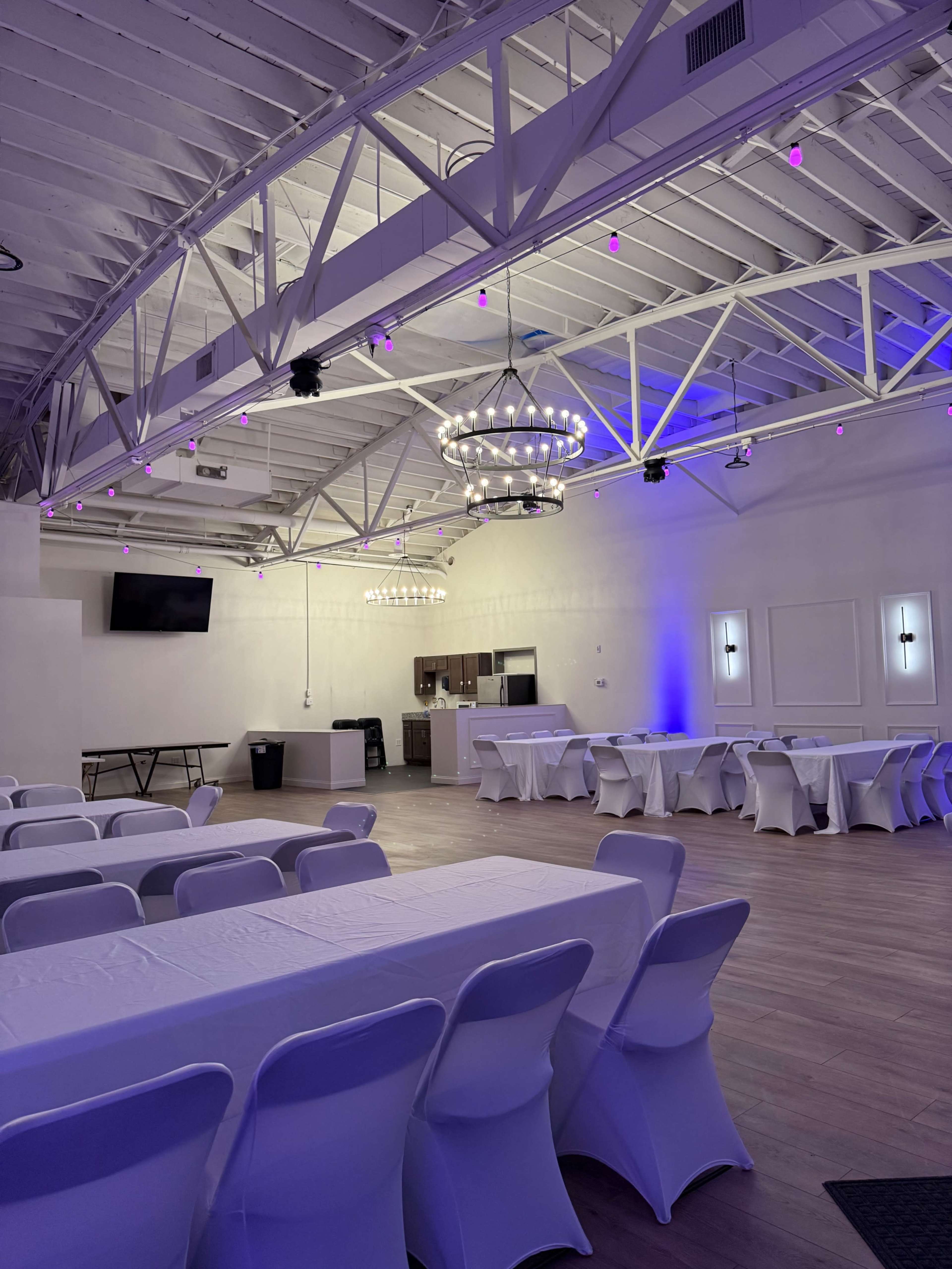 The image shows a spacious event hall with long tables covered in white cloths, a kitchen area in the background, and chandeliers hanging from the ceiling.