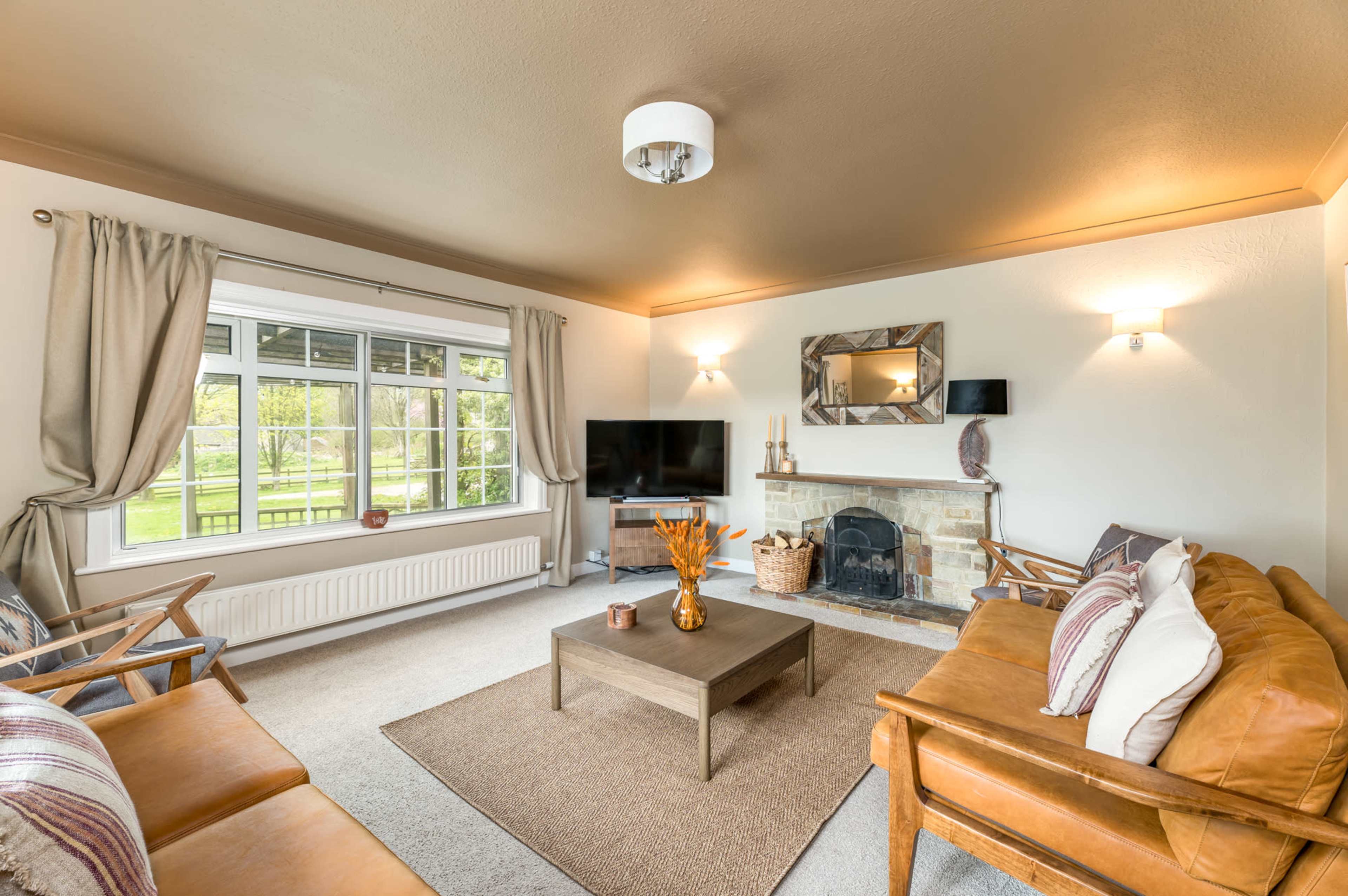 The image shows a cozy living room with a brown leather sofa, a wooden coffee table, and a large window overlooking a garden.