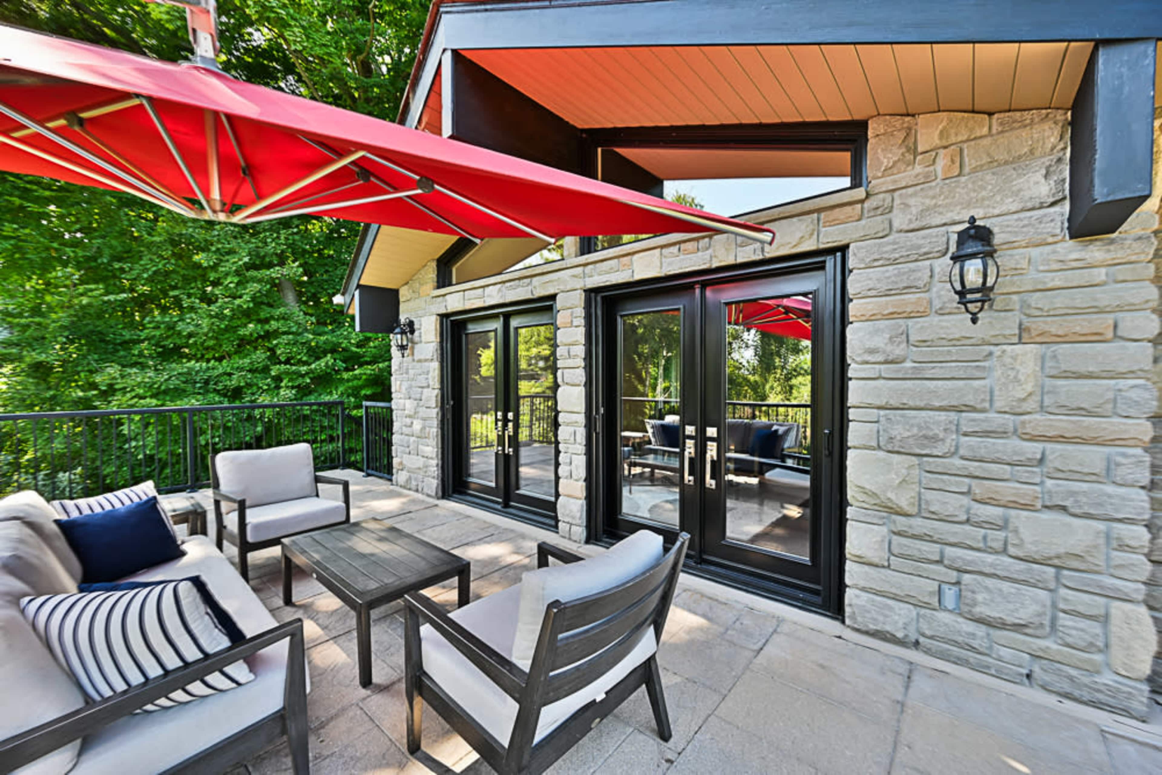 A patio with stone walls features a seating area arranged around a wooden table and is shaded by red umbrellas.