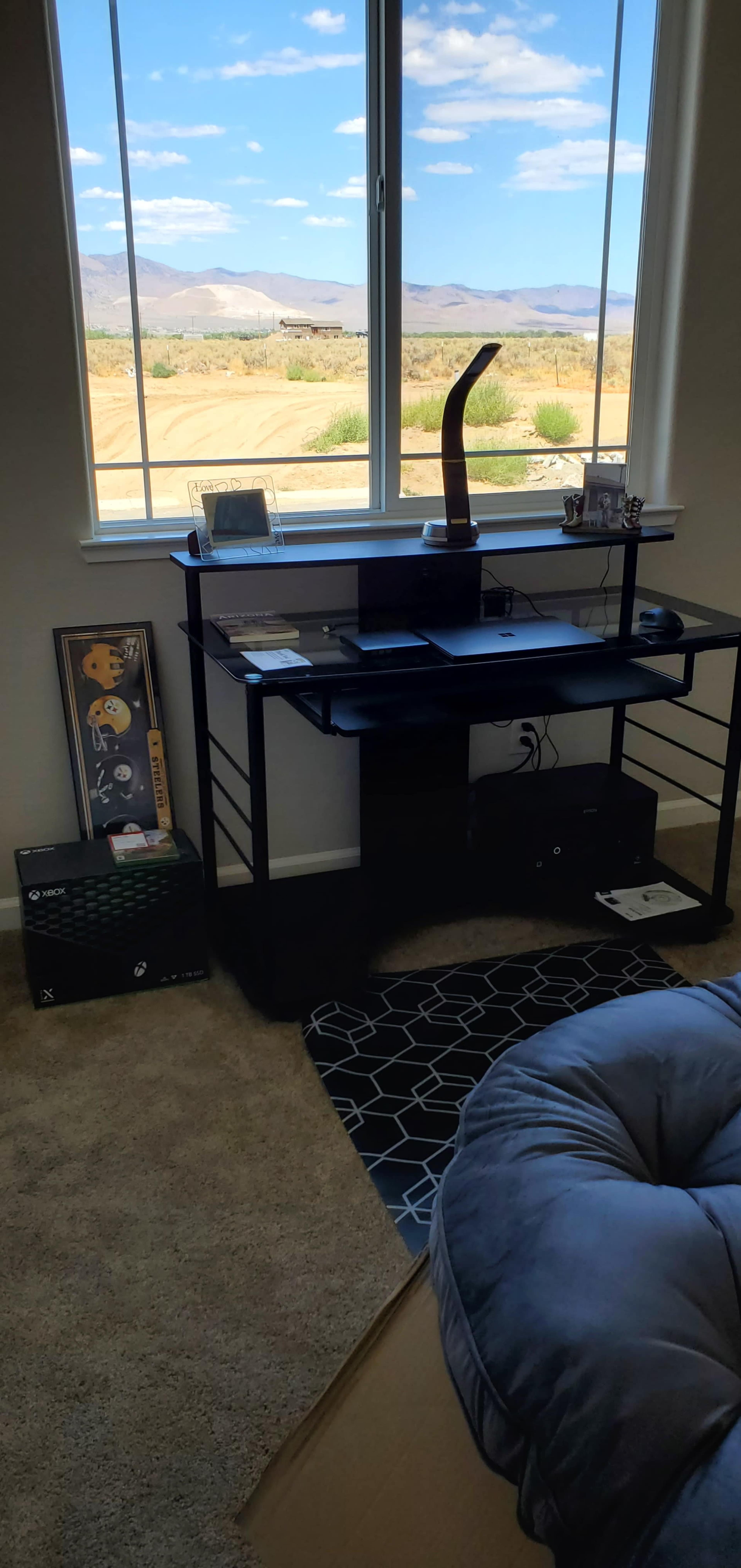 A black desk with a computer and decorative items sits near a large window overlooking a desert landscape with mountains in the distance.