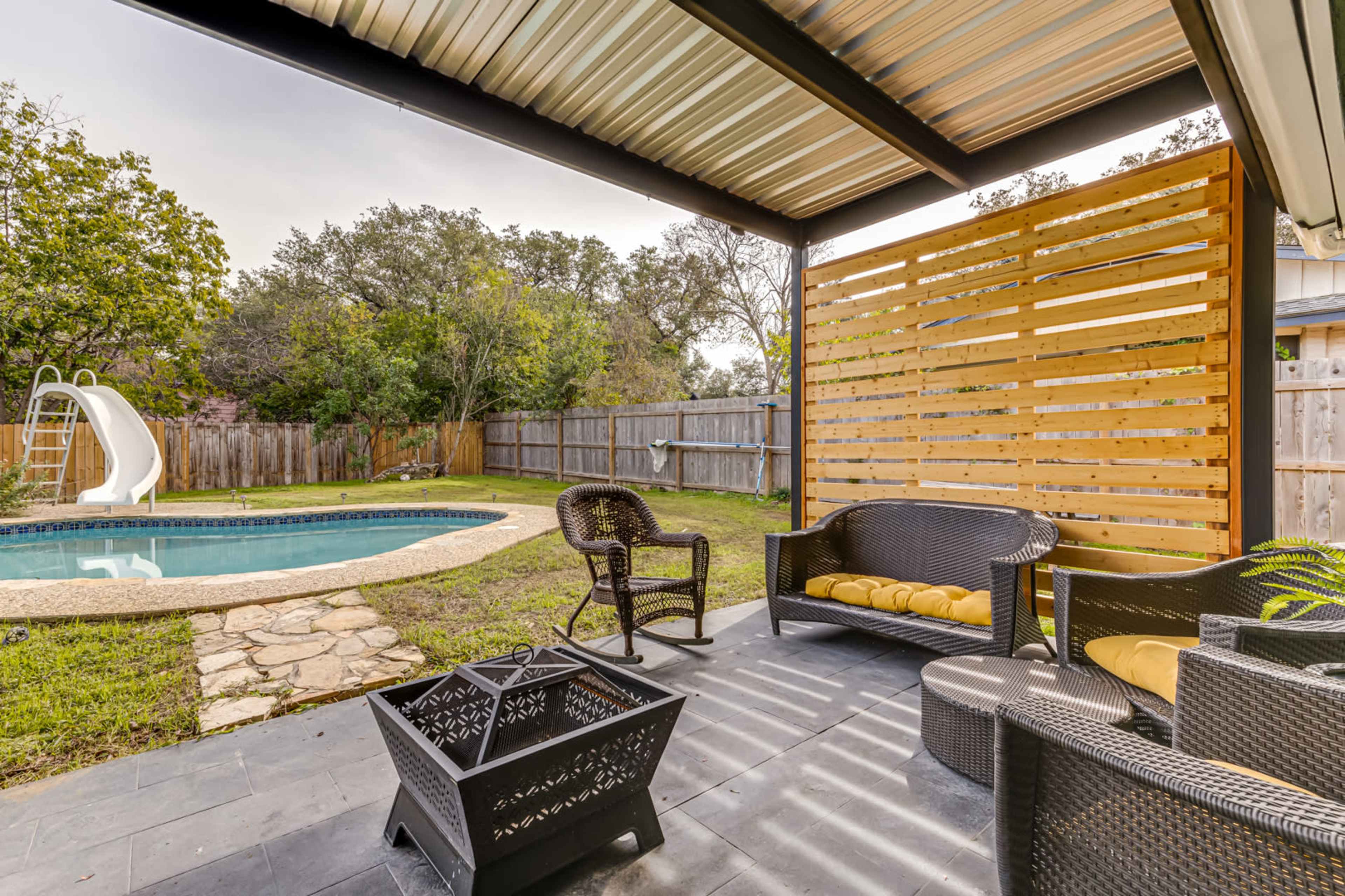 A backyard patio area with a pool, slide, and a seating arrangement featuring wicker furniture and a fire pit.