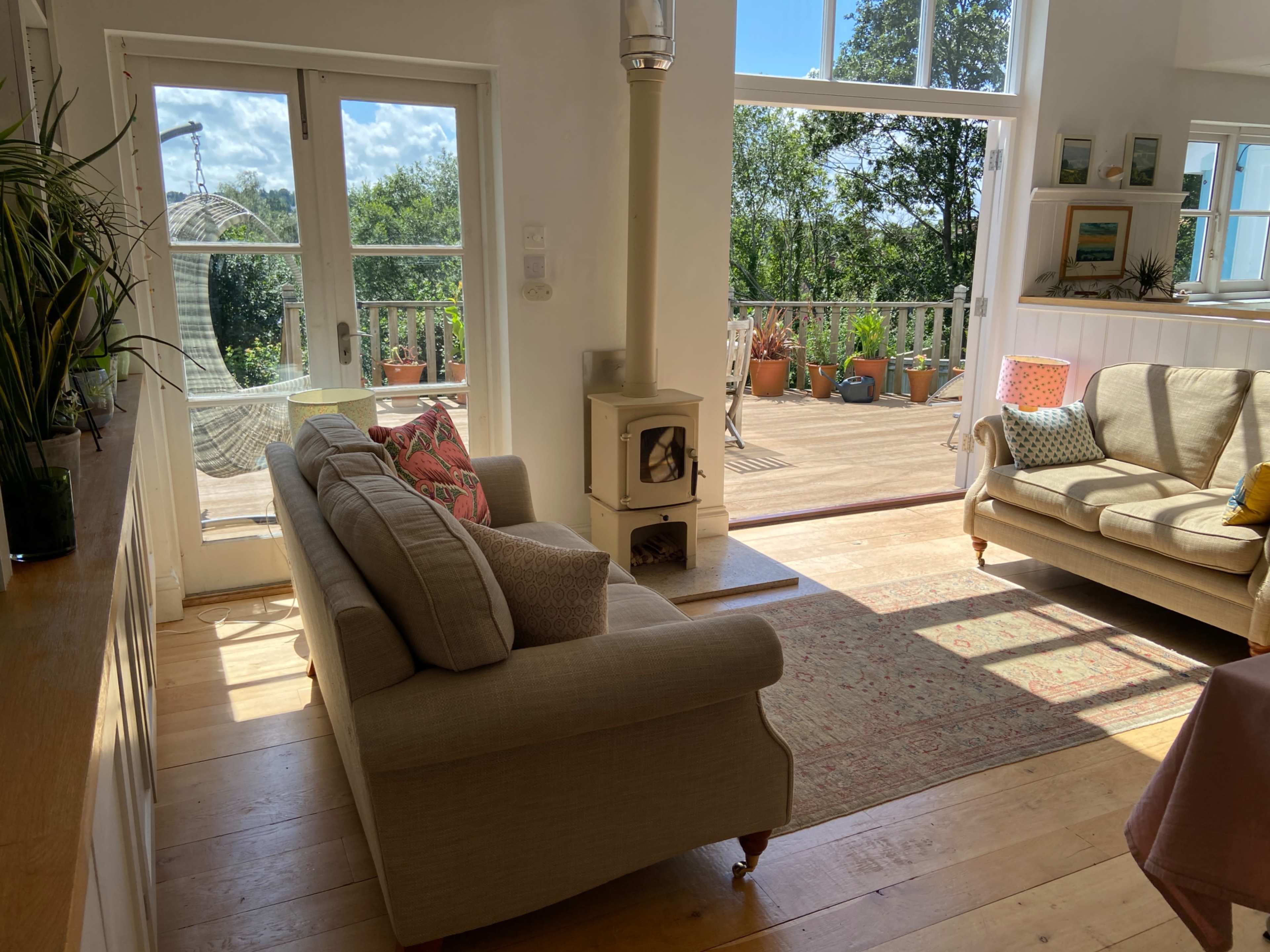 The image depicts a bright, modern living room with two sofas, a decorative rug, and large windows overlooking a porch and garden.