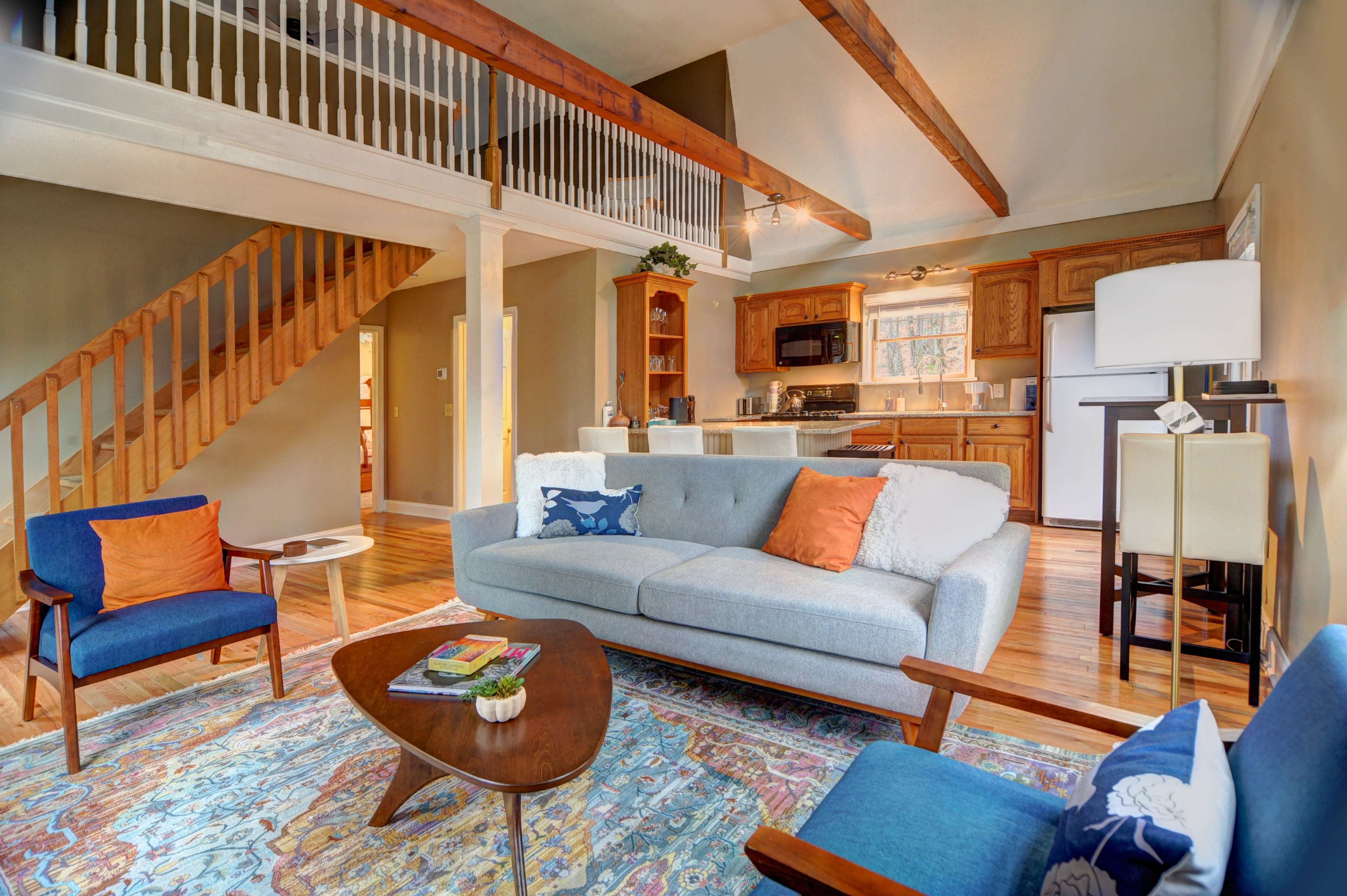 A bright living room with a gray couch, two blue chairs, a wooden coffee table, and a staircase leading to a loft area.