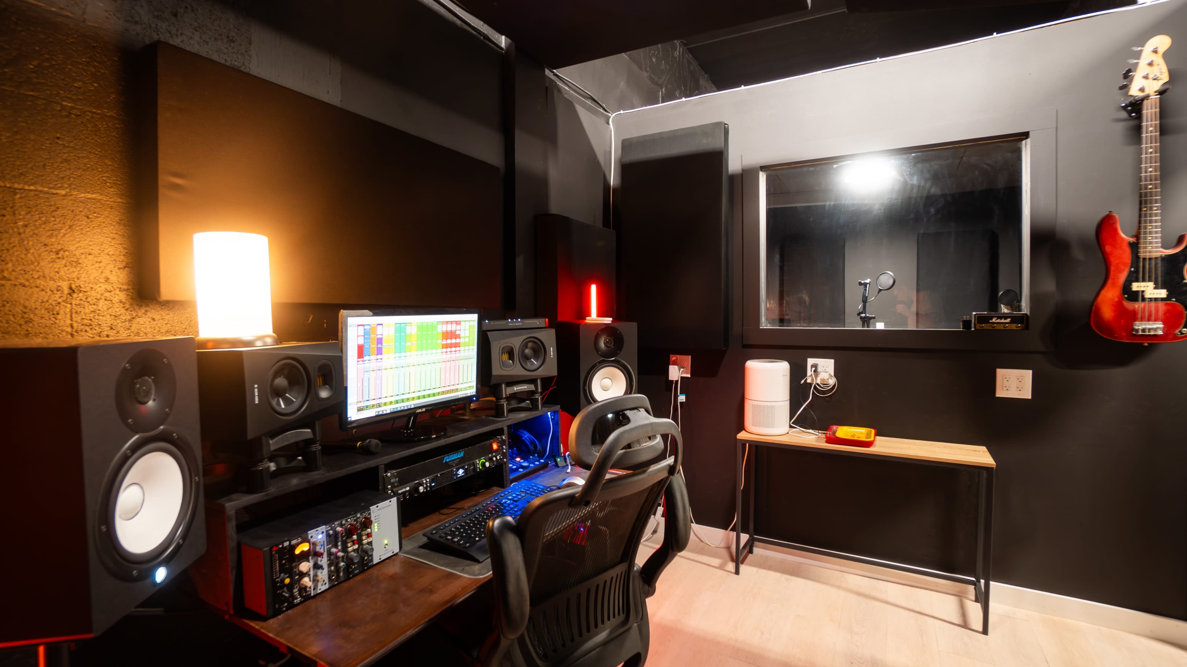 The image shows a modern music recording studio featuring a computer setup with audio monitors, a microphone booth visible through a glass window, and musical instruments mounted on the wall.