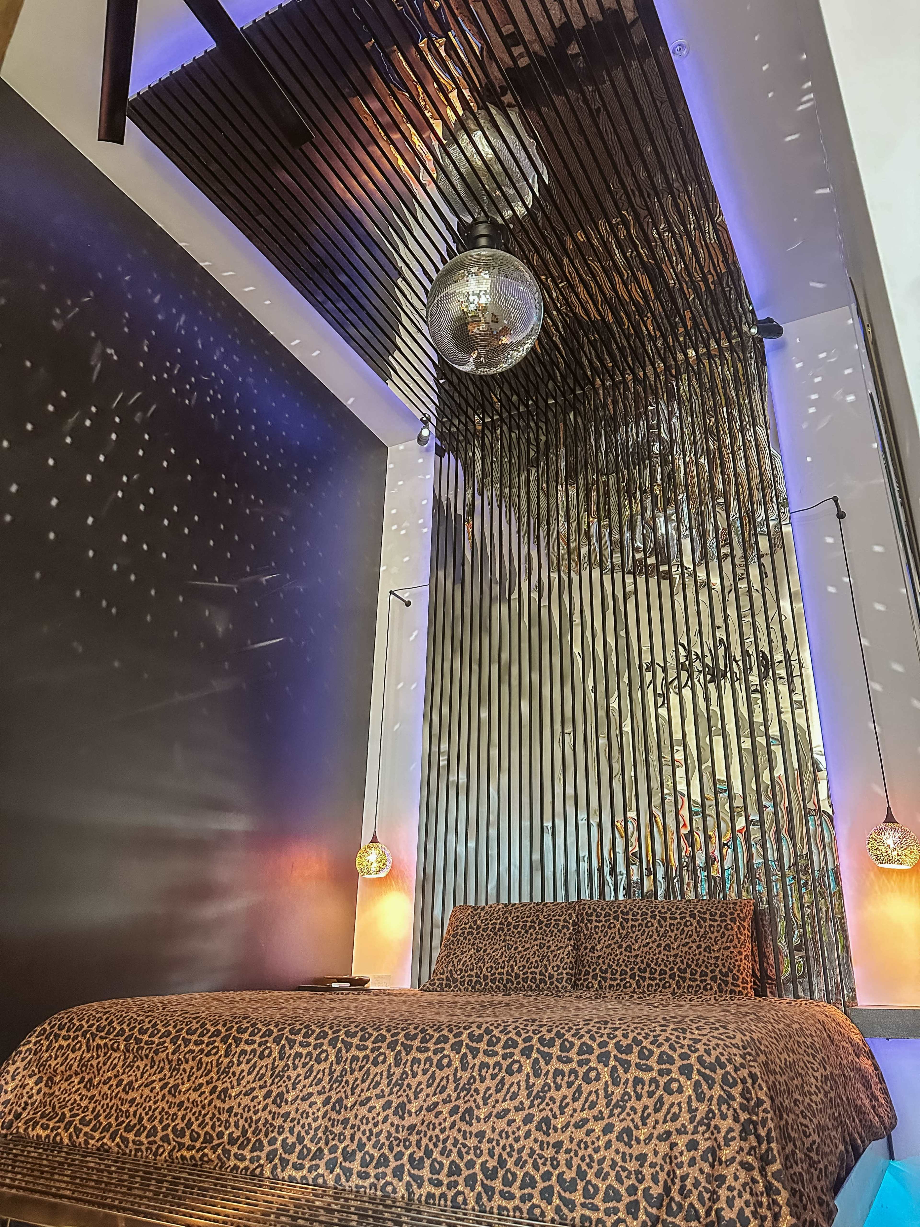 The image shows a modern bedroom featuring a leopard-print bedspread, a mirrored disco ball hanging from a wooden ceiling, and ambient lighting illuminating the walls.