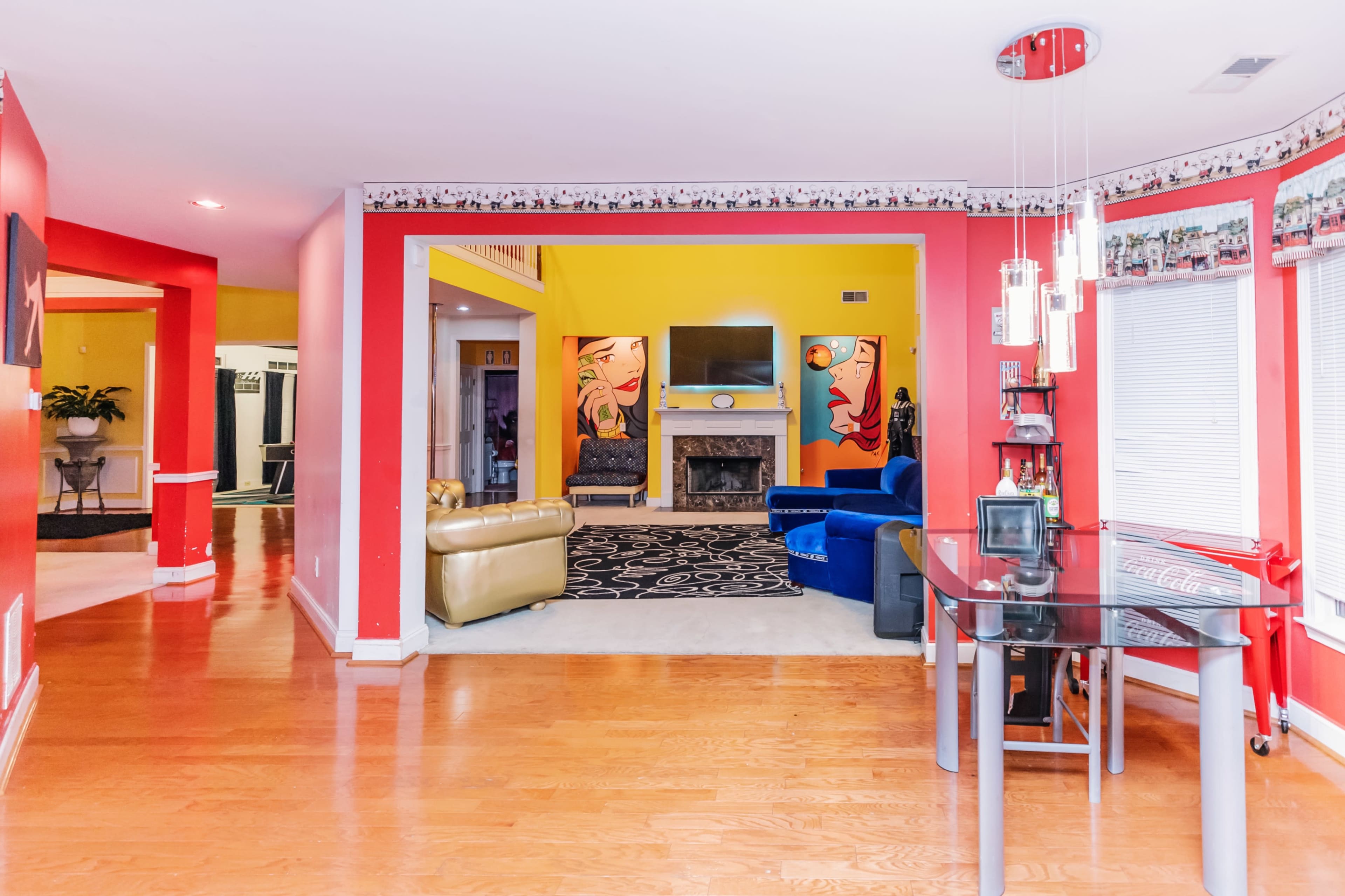 The image shows a brightly colored living space with a pink and yellow color scheme, featuring a mix of furniture including a yellow sofa, a blue armchair, and a glass dining table.