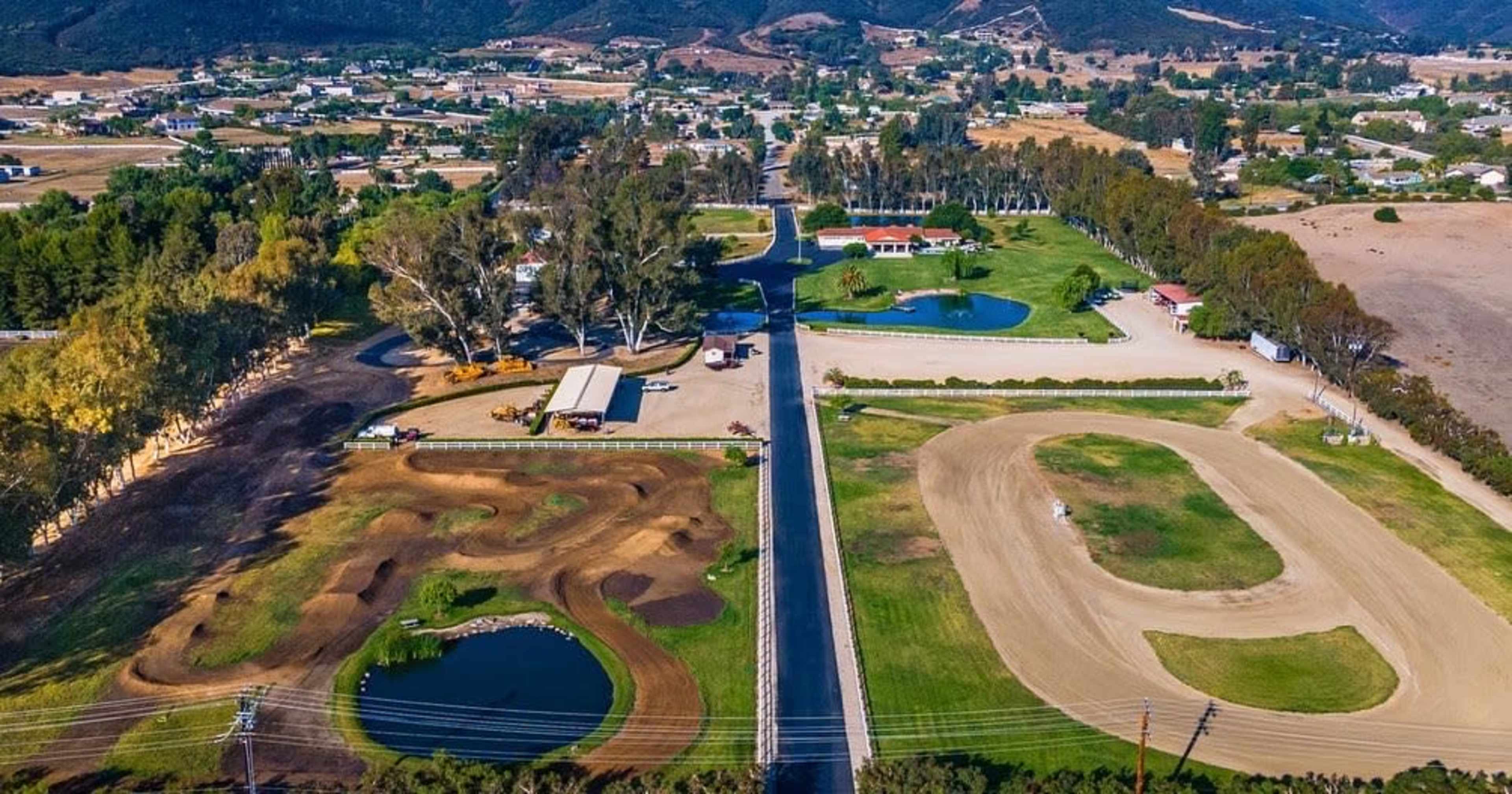 An aerial view of a rural area featuring a horse track, a motocross track, and several ponds surrounded by trees and buildings.