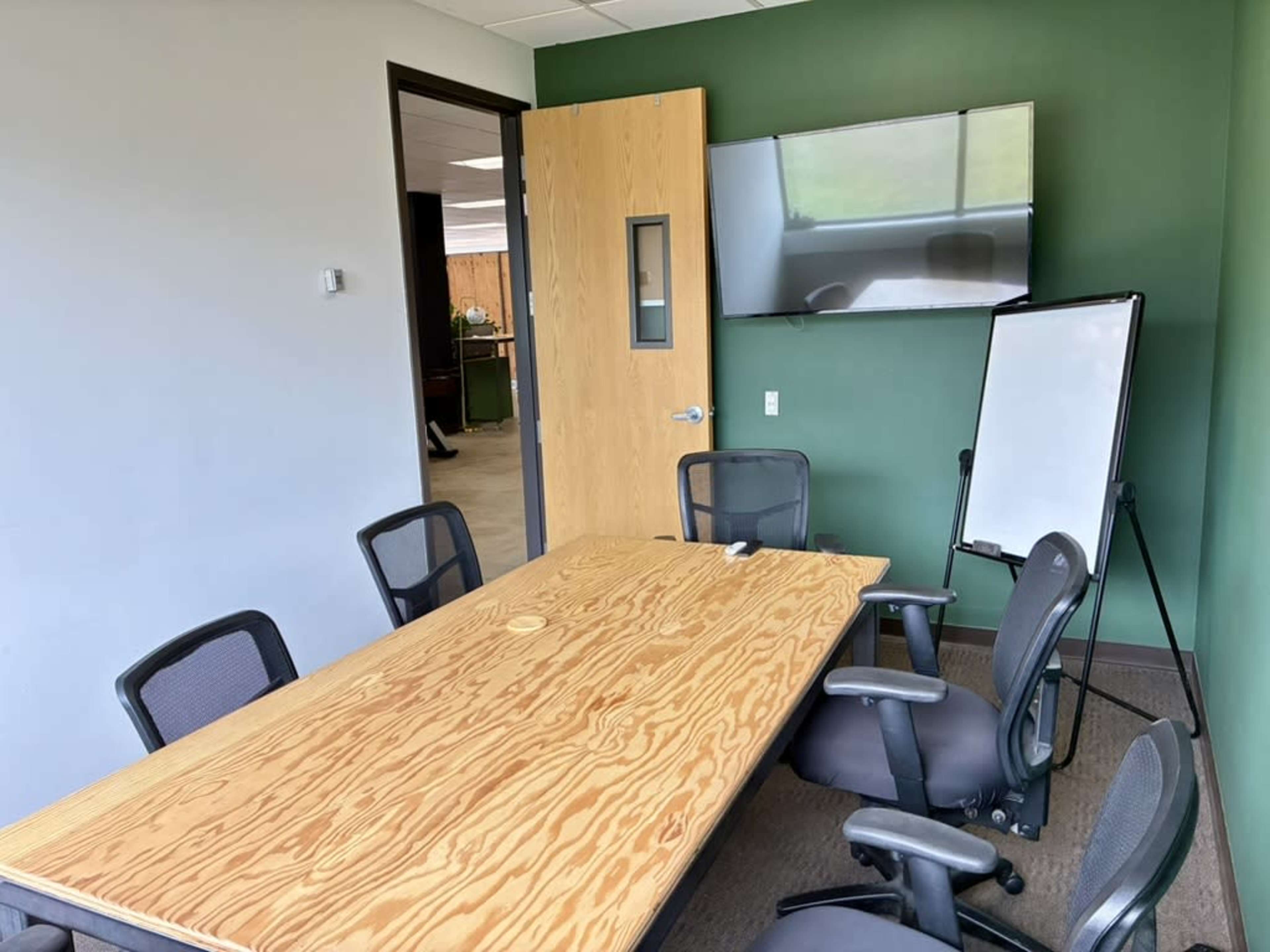 The image shows a conference room featuring a wooden table, several black chairs, a whiteboard on a stand, and a wall-mounted television.